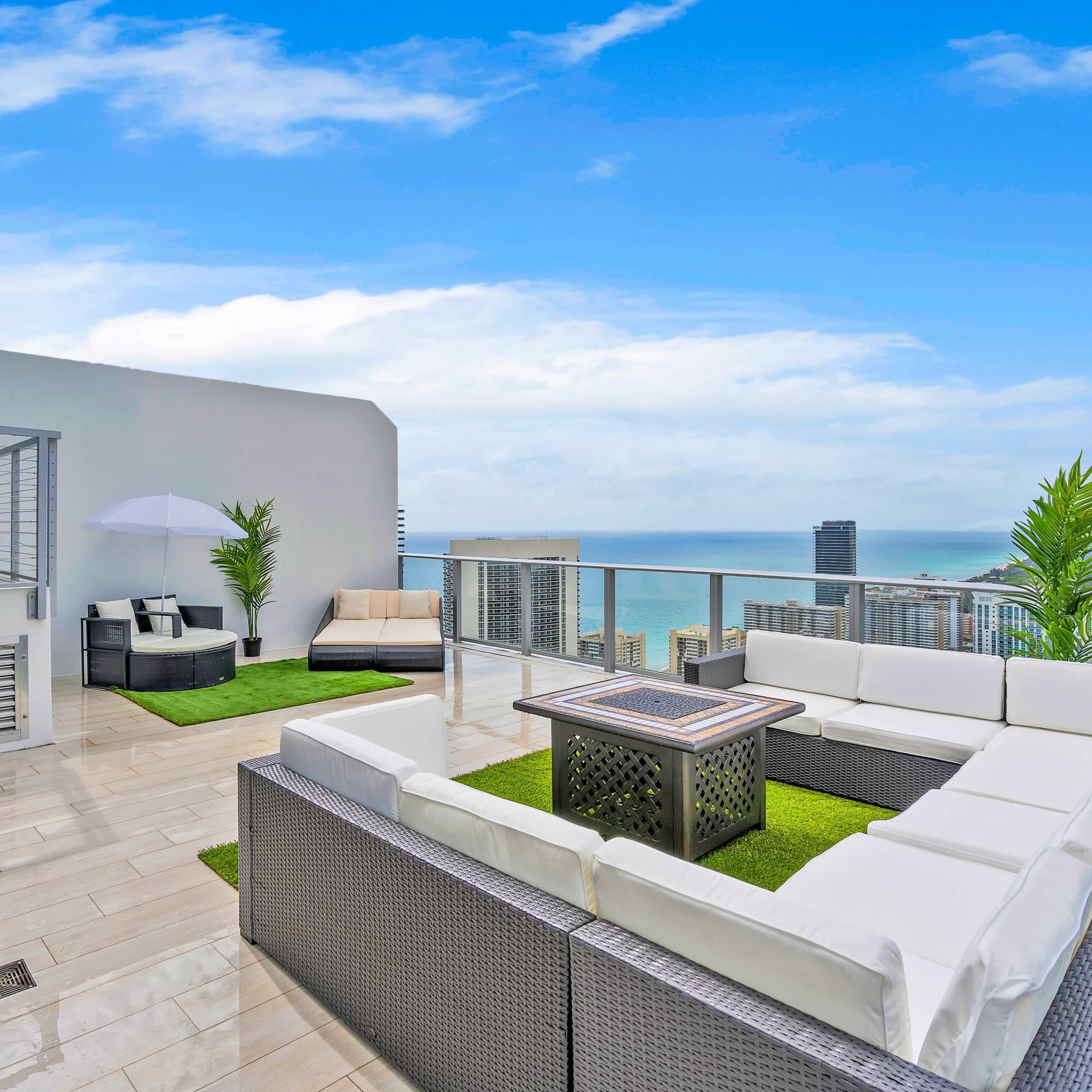 A modern rooftop terrace featuring a seating area with white outdoor furniture, a coffee table, and a view of the ocean and city skyline.
