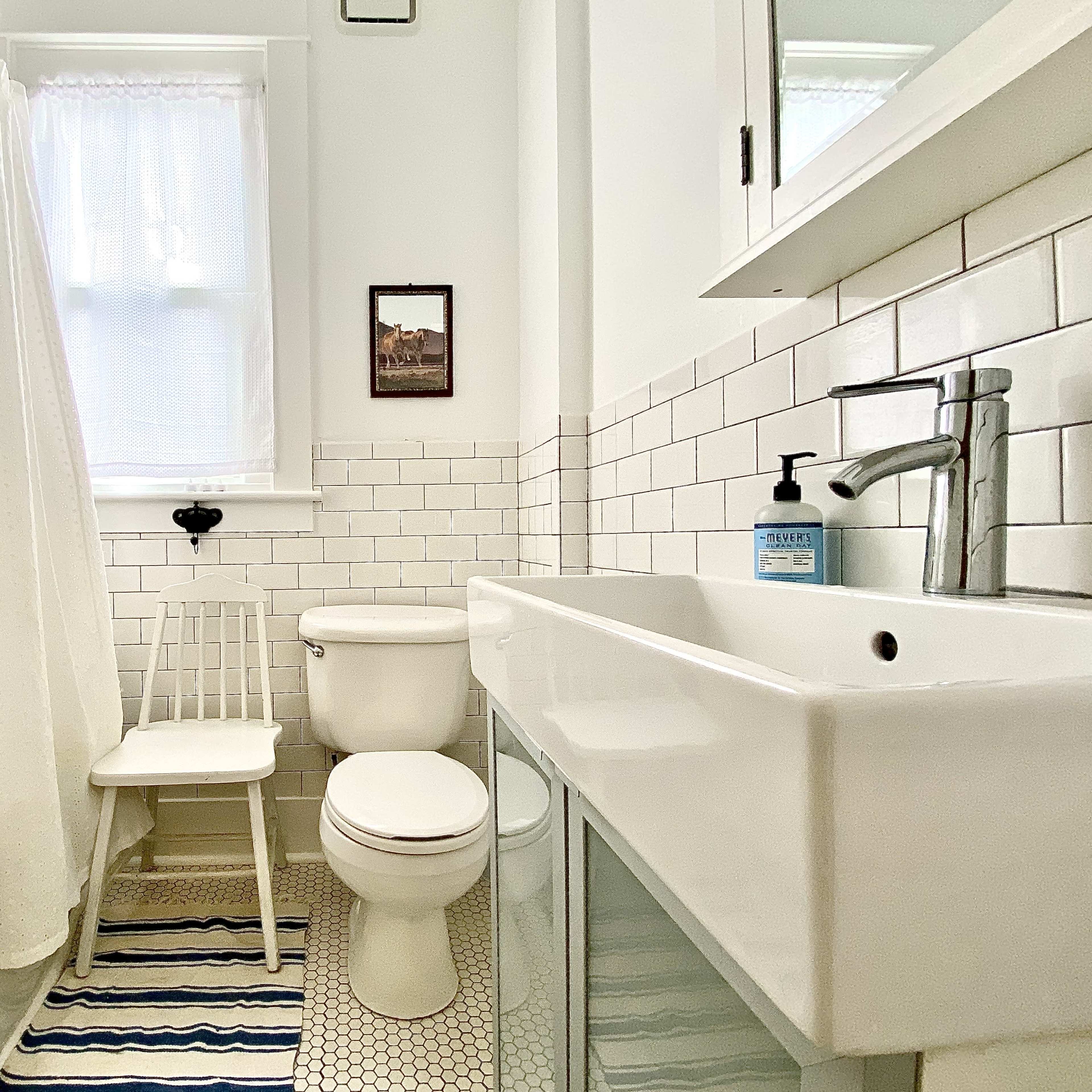 A bright bathroom featuring a white tiled shower with a curtain, a toilet, a sink with a mirror above it, and a chair beside the tub.