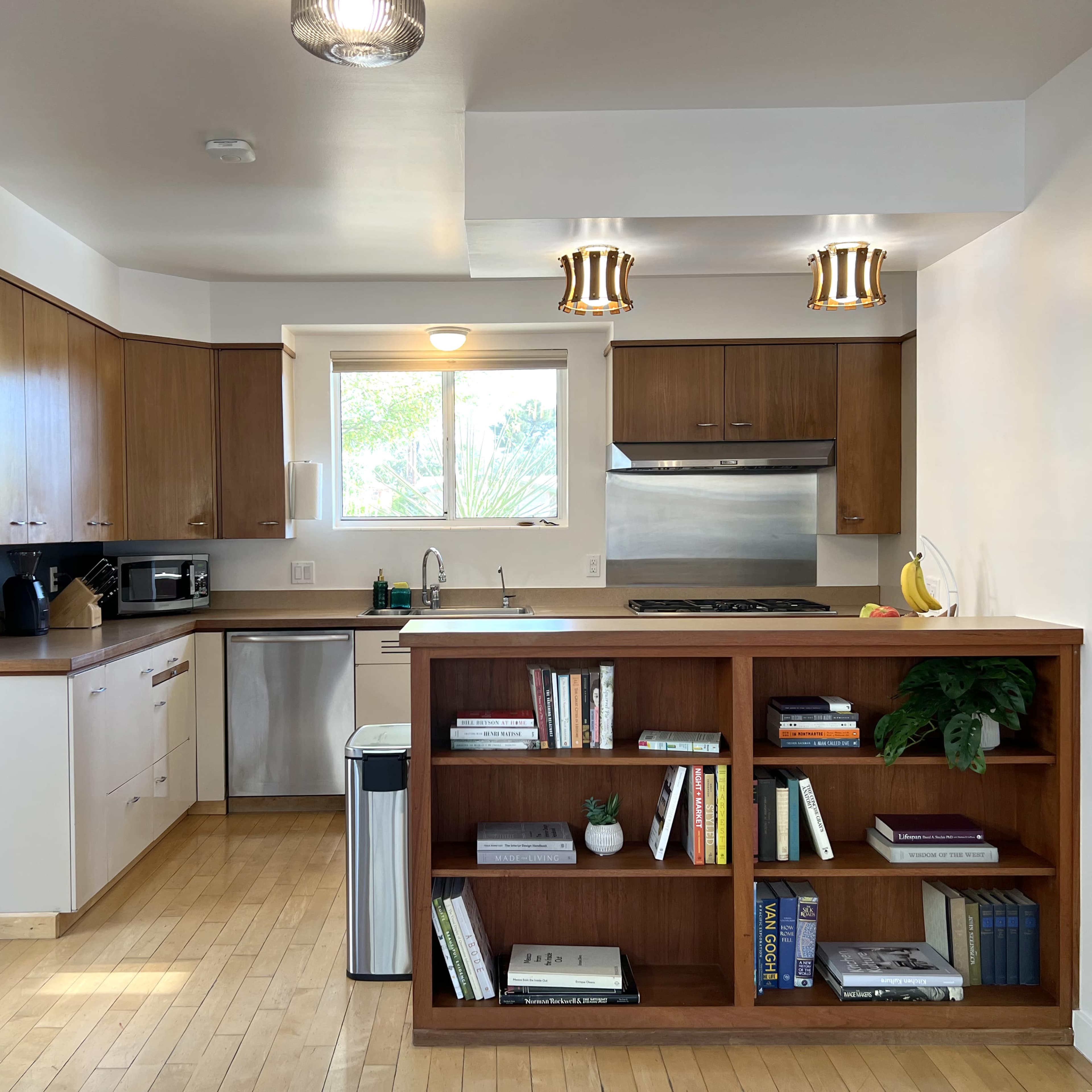 A modern kitchen features wooden cabinetry, stainless steel appliances, and a bookshelf with various books and a plant.