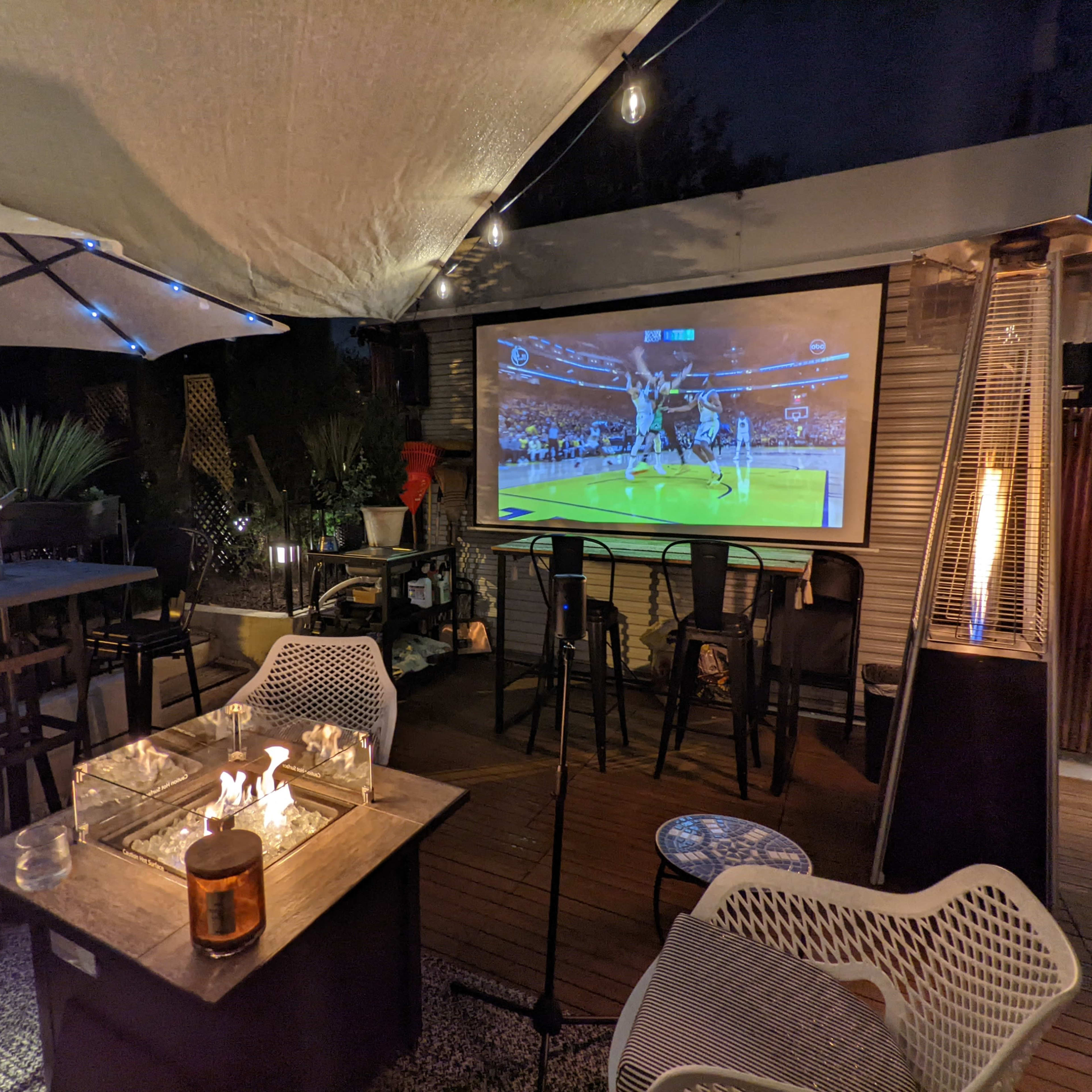 An outdoor seating area features a large screen displaying a basketball game, surrounded by plants, bar stools, and patio lighting.
