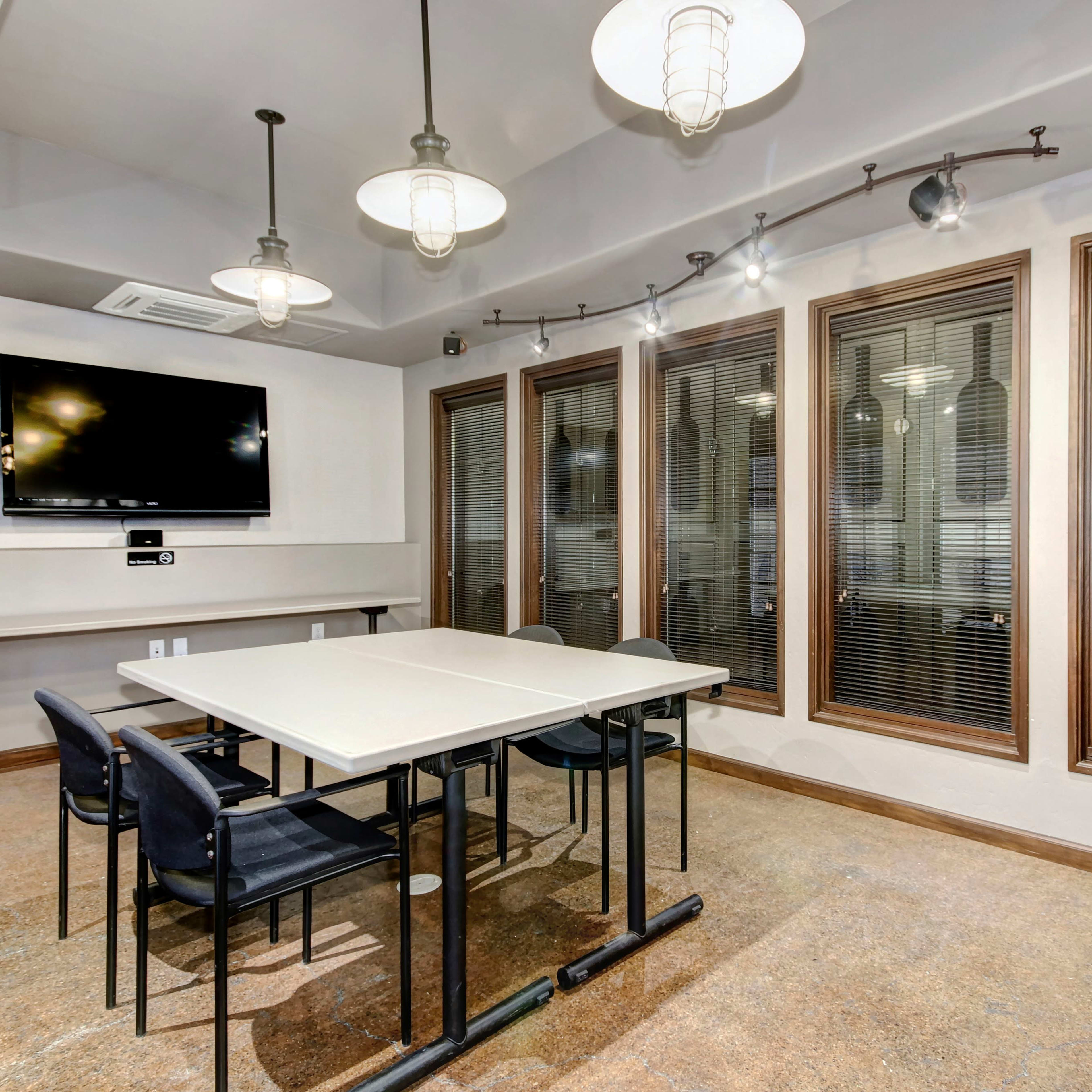 A conference room features a rectangular table surrounded by black chairs, a wall-mounted screen, and large windows with blinds.