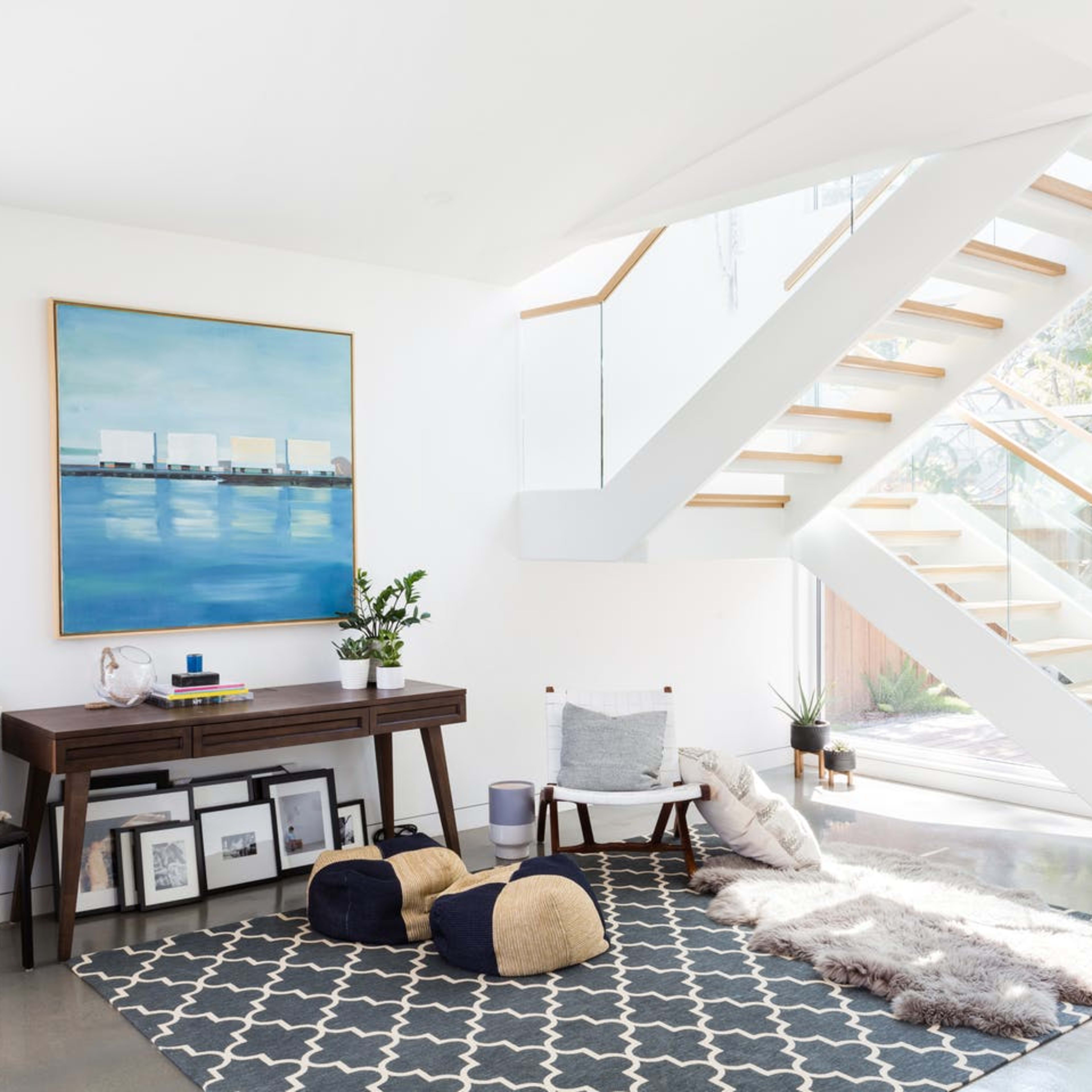 A modern interior featuring a staircase, a desk with a plant, and a seating area with poufs on a patterned rug.