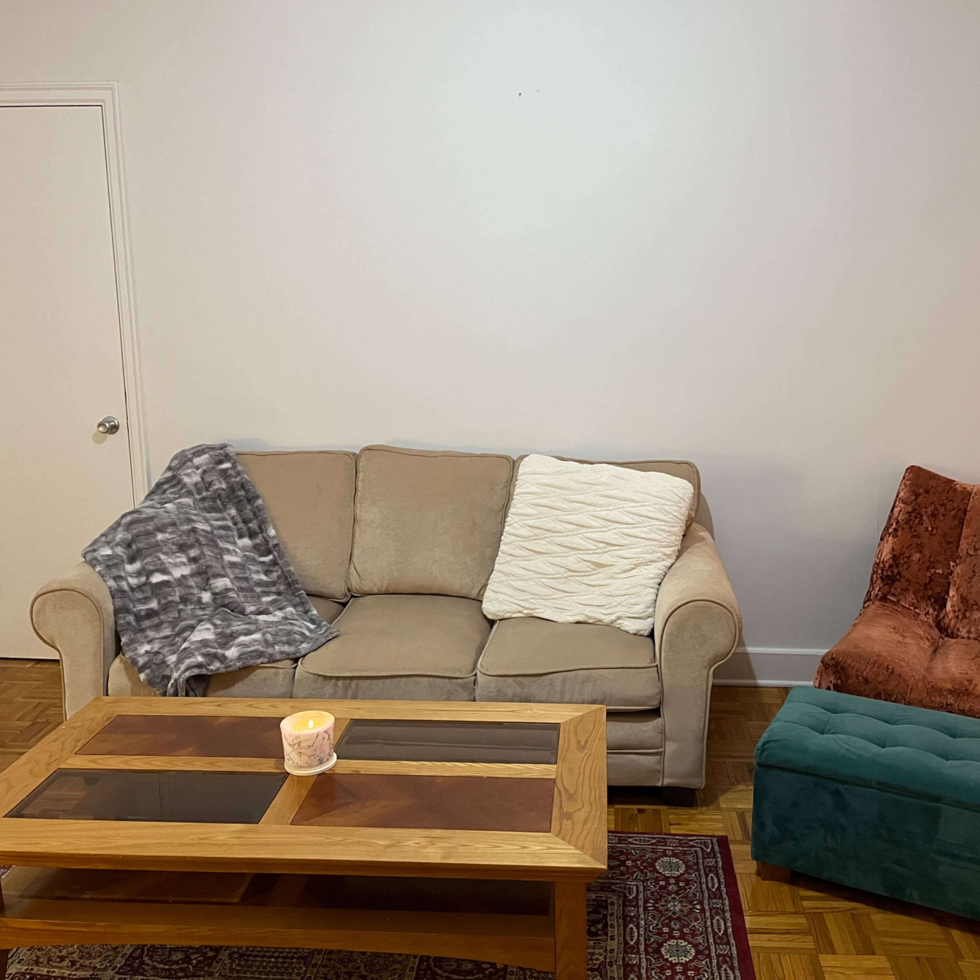 A living room with a beige couch, an orange chair, a wooden coffee table, and a candle on a patterned rug.
