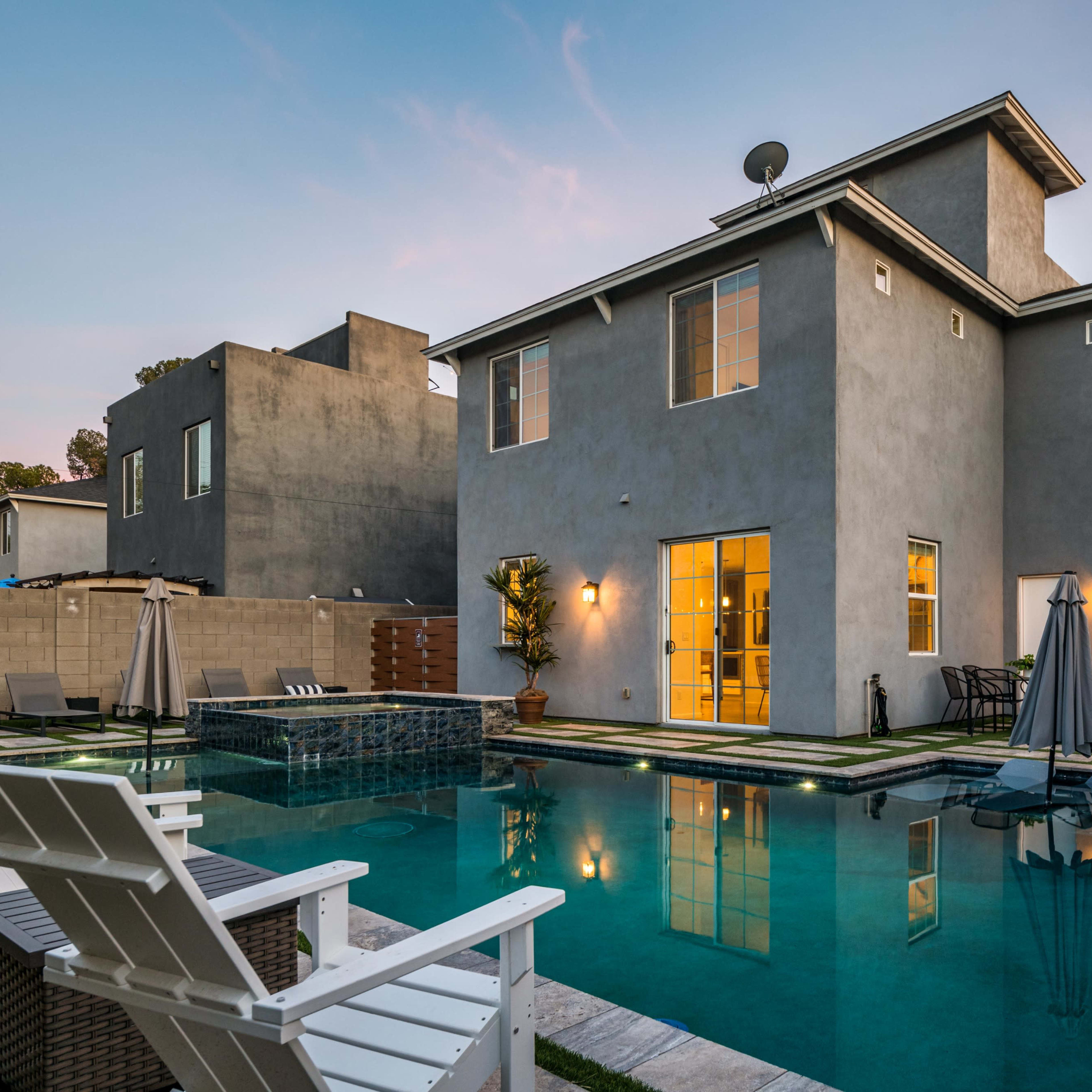 A modern home with a backyard pool, loungers, and a patio area, surrounded by greenery at twilight.