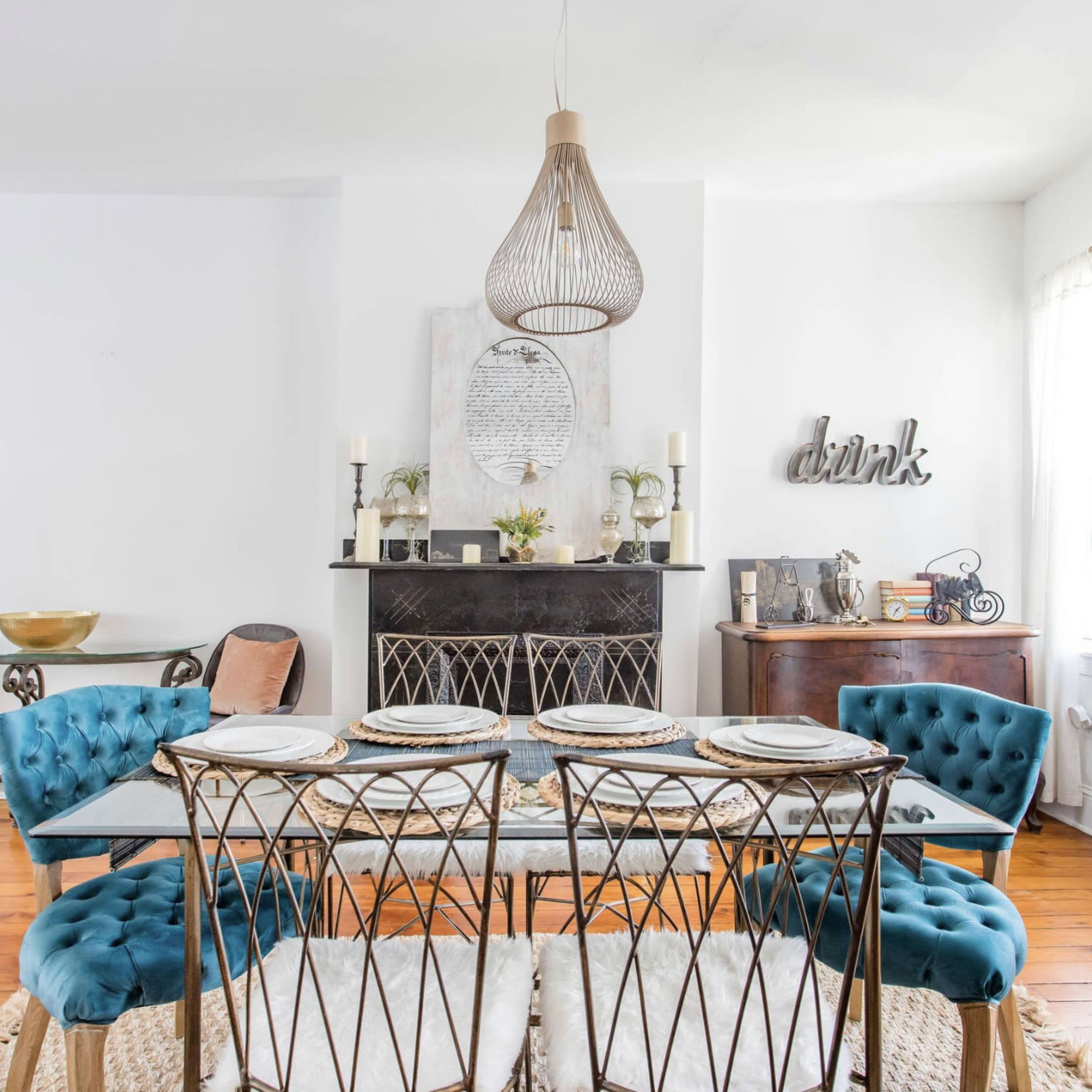 A dining area features a glass table with tufted blue chairs, a dark fireplace, and decorative elements on the walls.