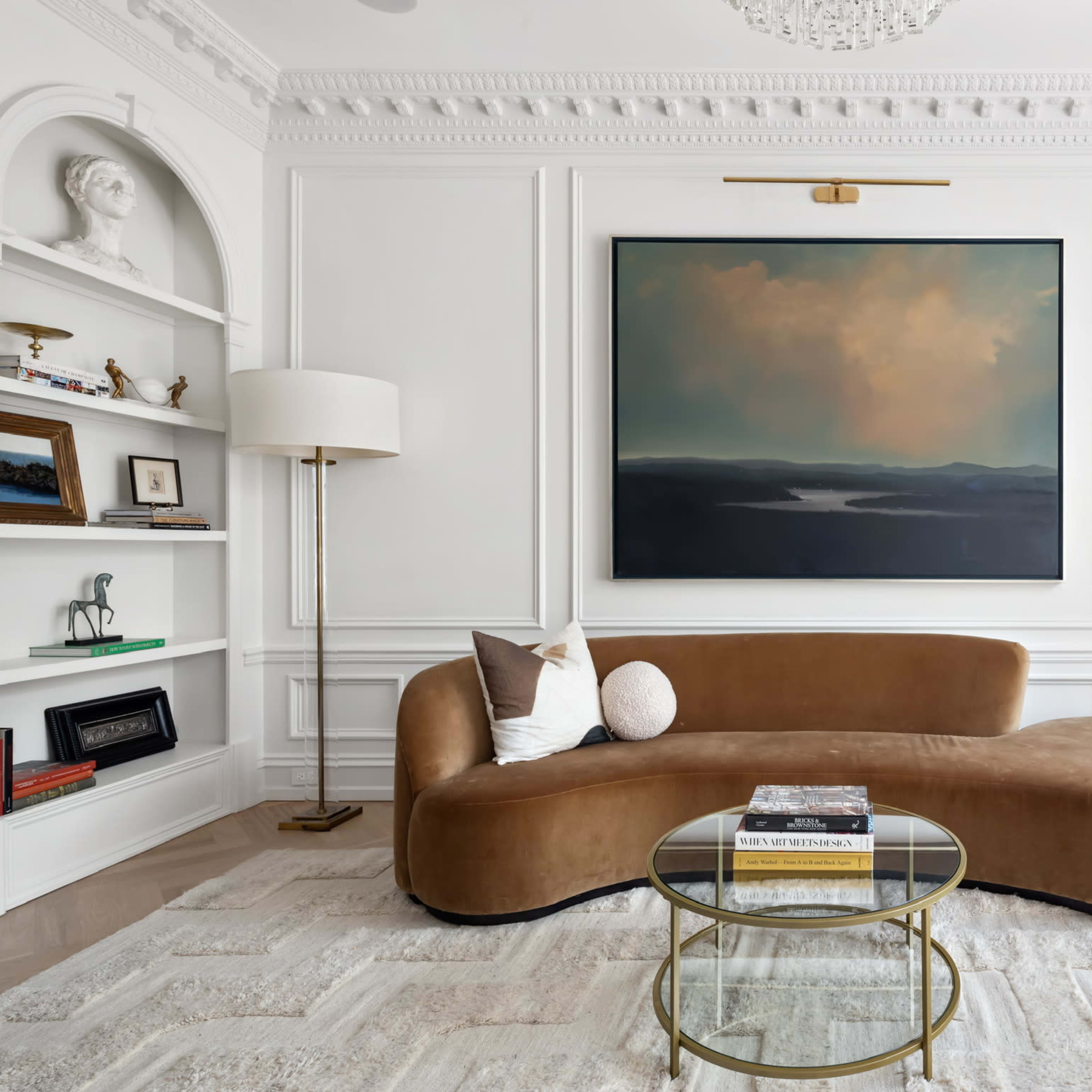 A modern living room featuring a curved brown sofa, a glass coffee table, two floor lamps, and a large painting mounted on a white paneled wall.