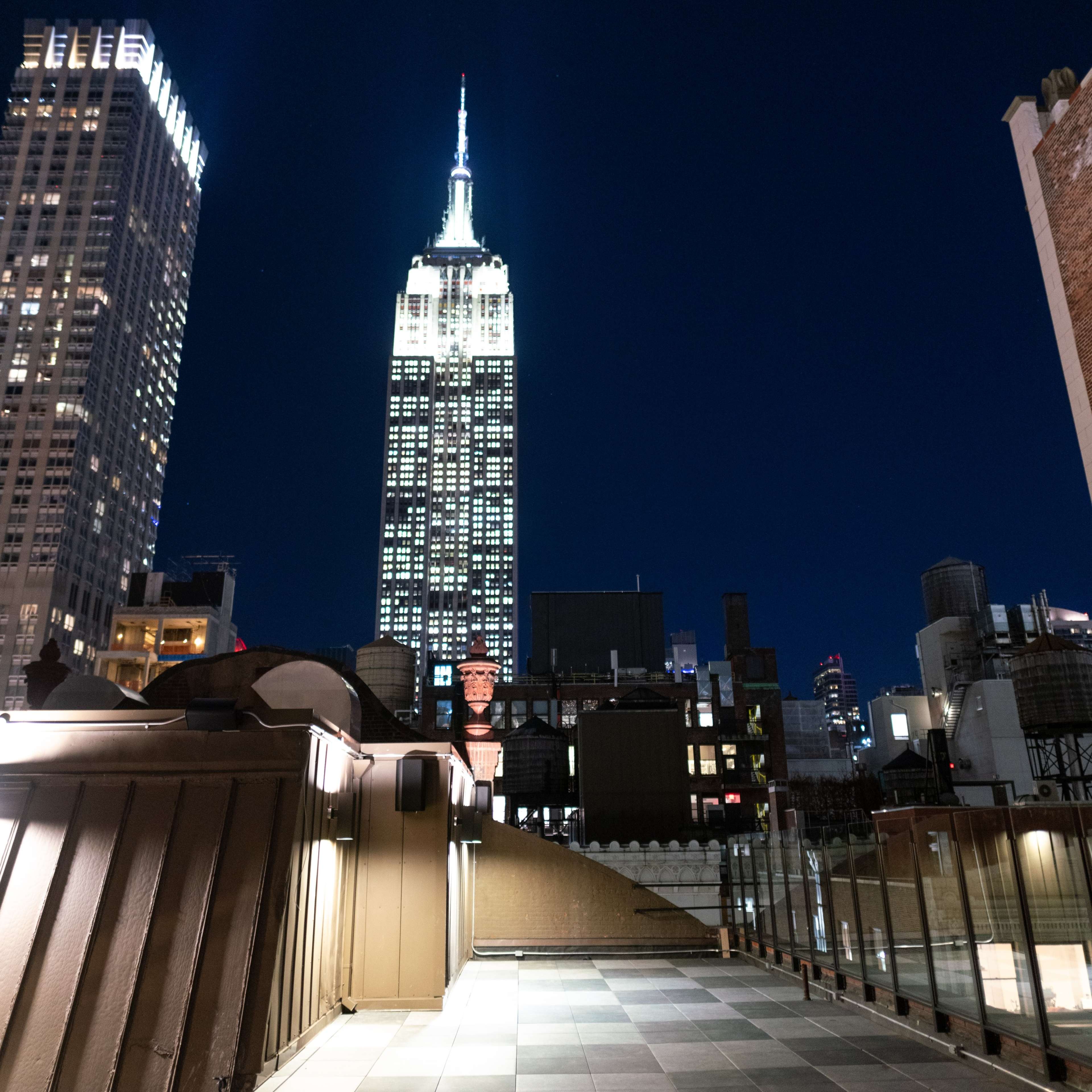 A rooftop view at night featuring the brightly lit Empire State Building surrounded by city buildings.
