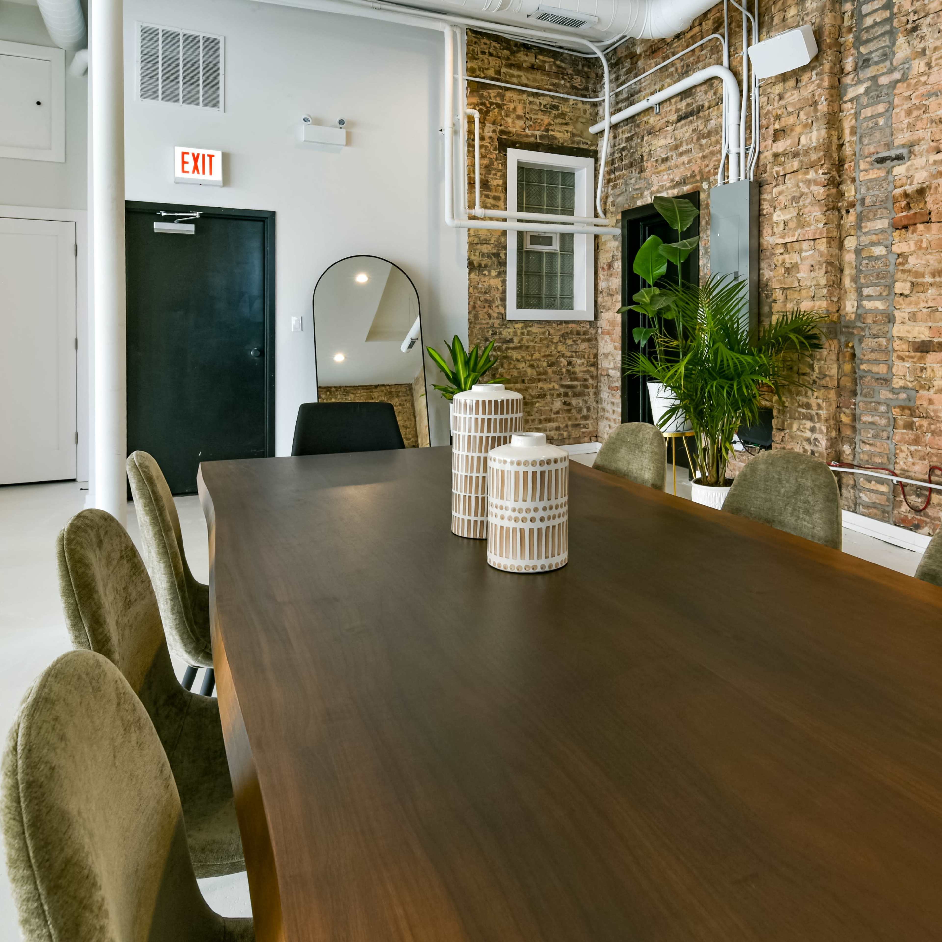A long wooden dining table is set in a room with exposed brick walls and modern furnishings, featuring two decorative vases and a large mirror.