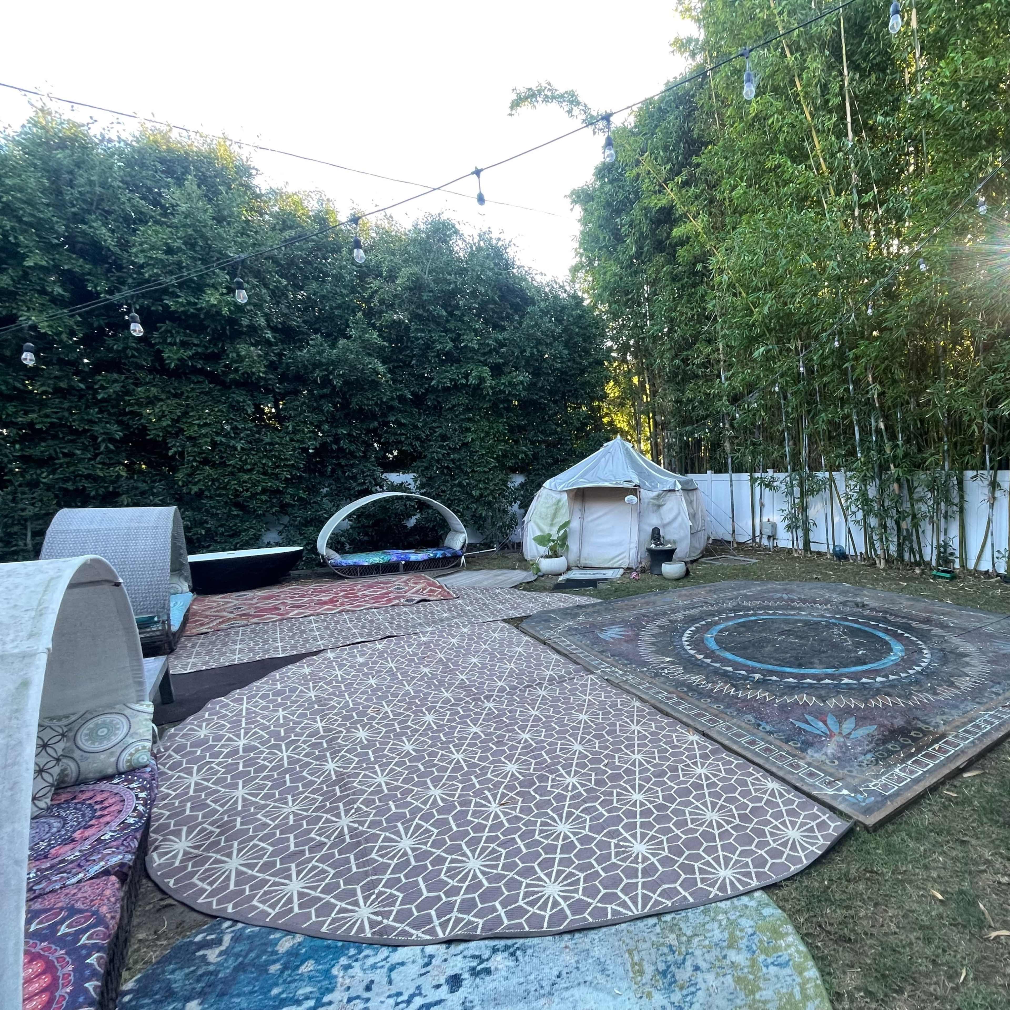 The scene features a backyard with several decorative rugs, a small tent structure, and a backdrop of lush greenery and bamboo.