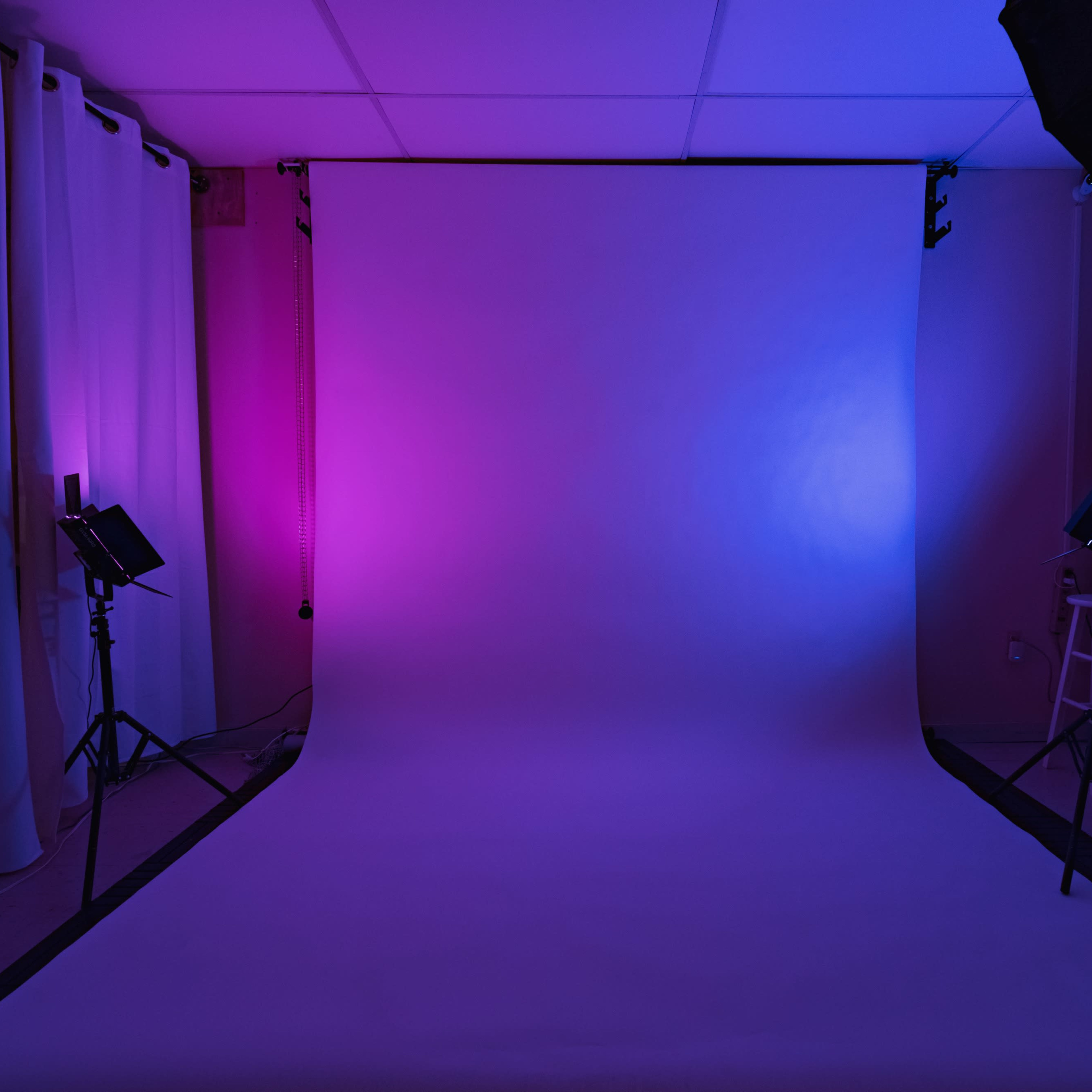 A photography studio with a backdrop illuminated by purple and blue lights, flanked by white curtains and lighting equipment.