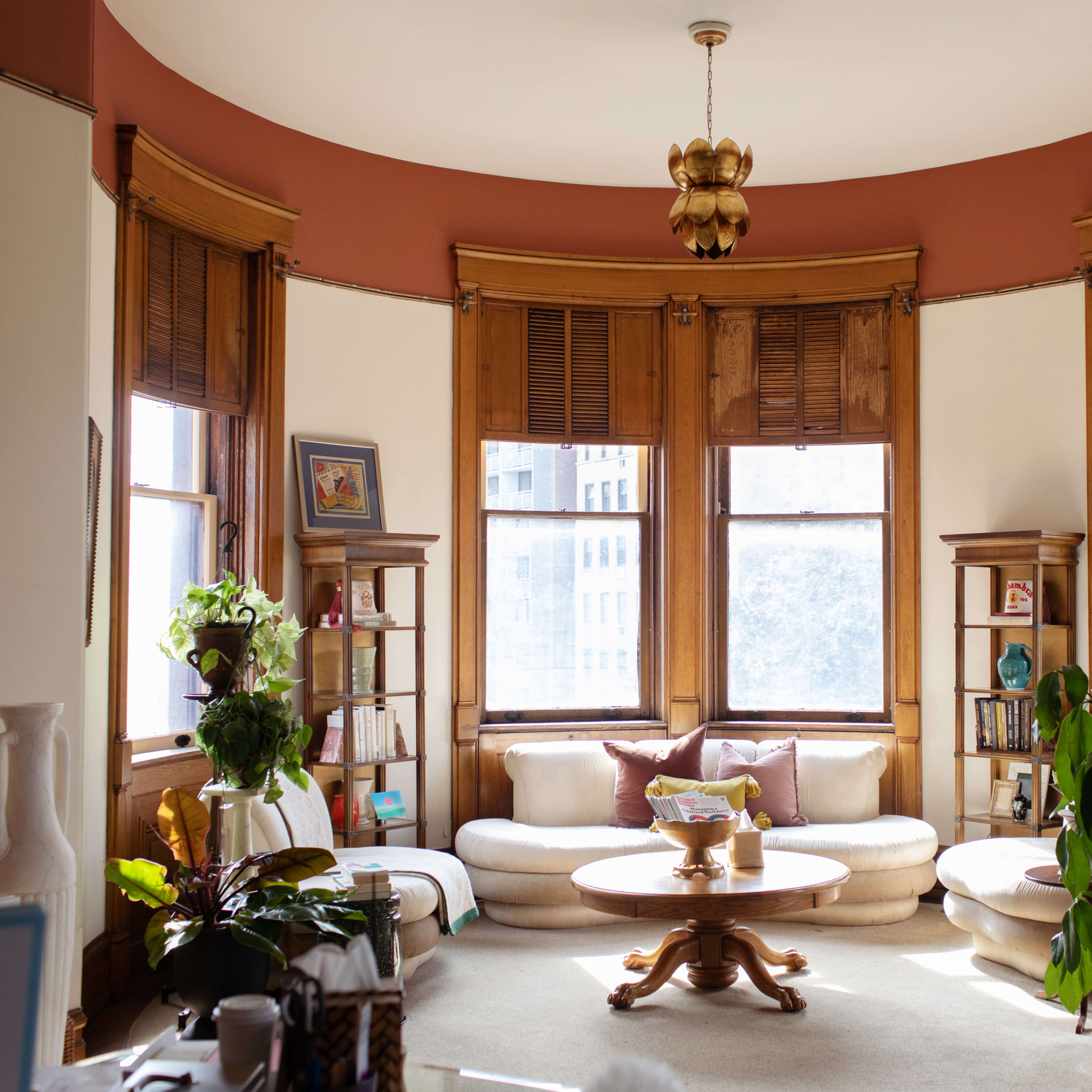 A sunlit living room features curved windows, a round coffee table, and light-colored seating arranged around a central area with bookshelves and plants.