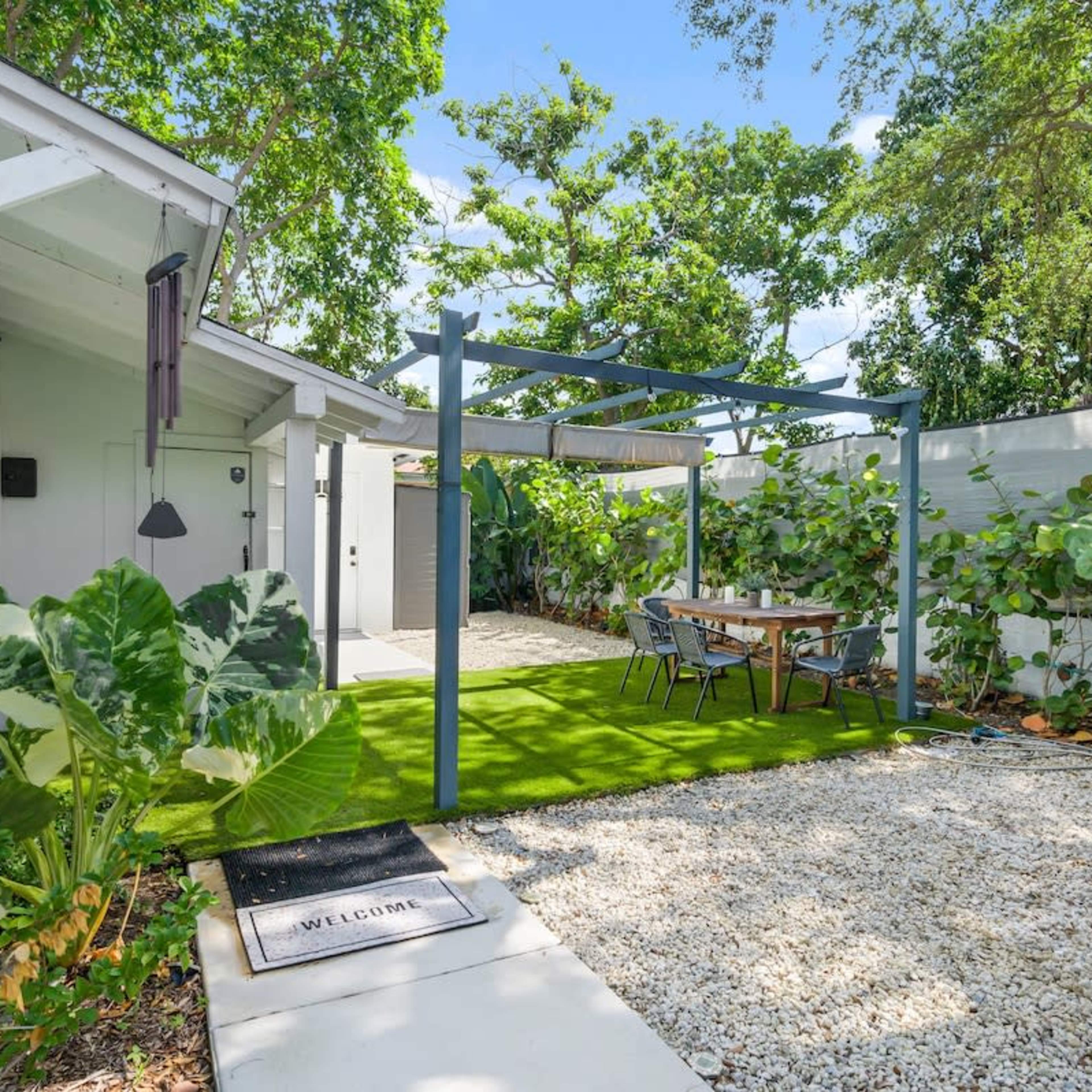 A small outdoor seating area with a table under a pergola, surrounded by greenery and gravel pathways next to a house.