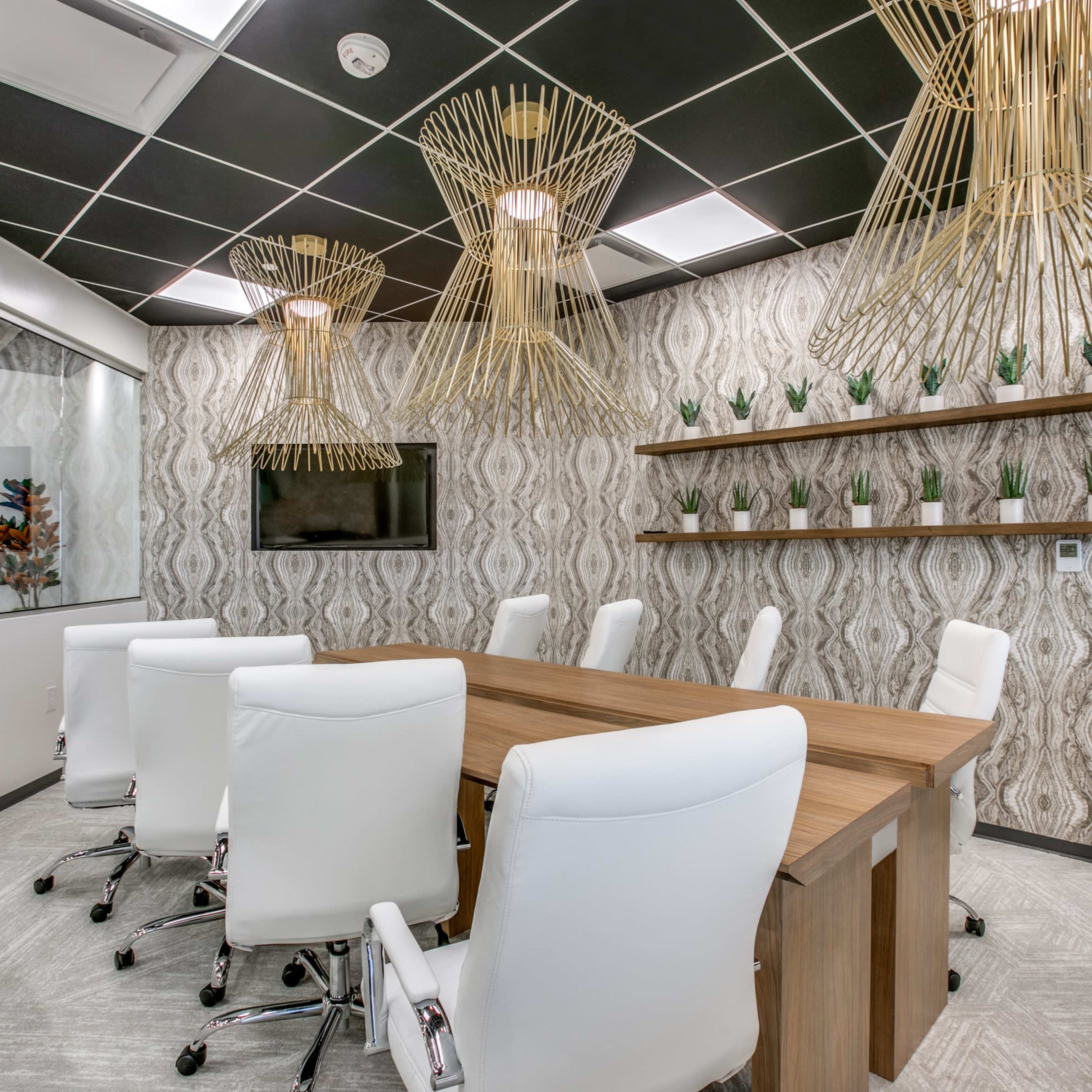 A modern conference room features a large wooden table surrounded by white leather chairs, with decorative hanging lights and potted plants on the walls.