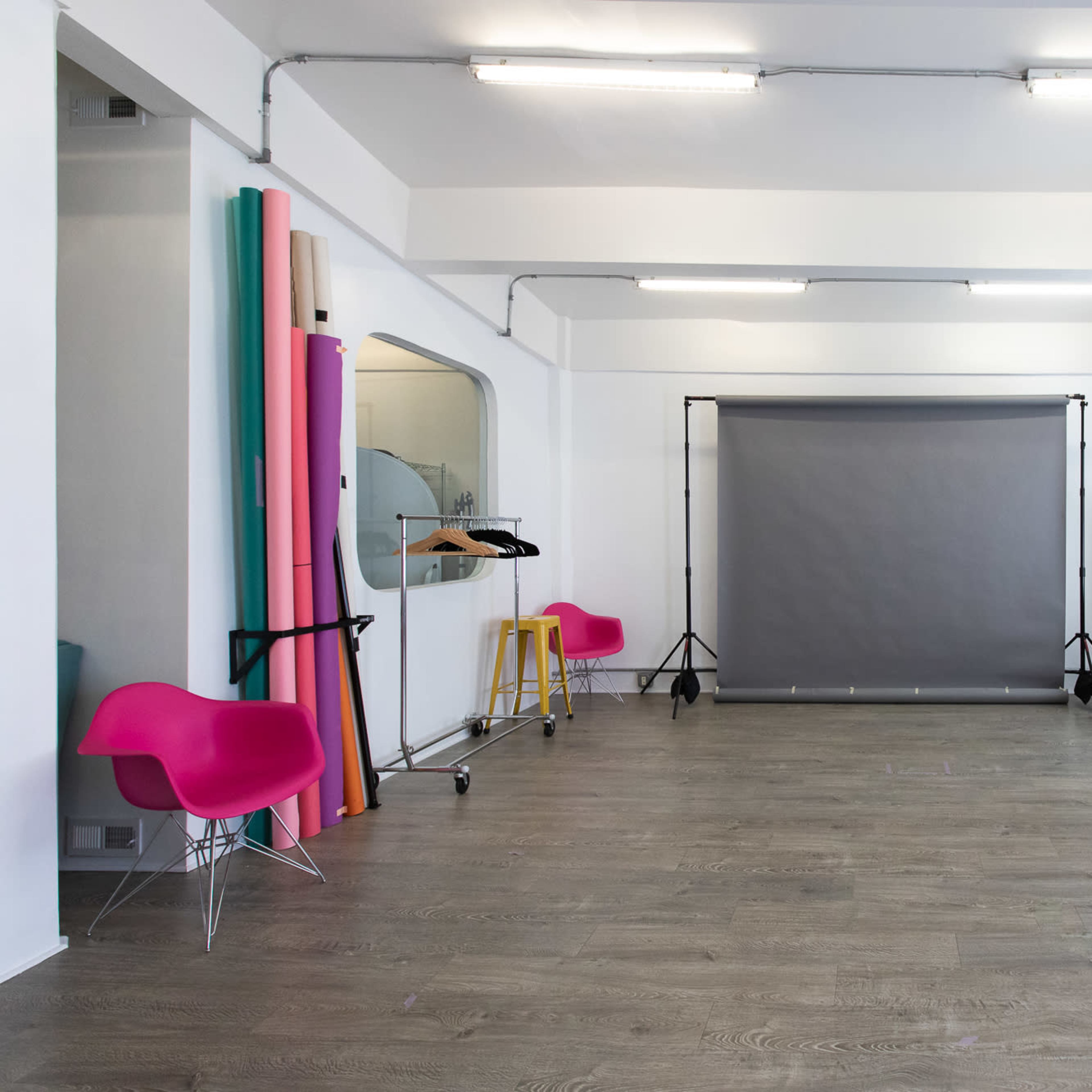 A spacious studio with a gray backdrop, colorful rolls of backdrop material, mirrors, and several chairs arranged in the room.
