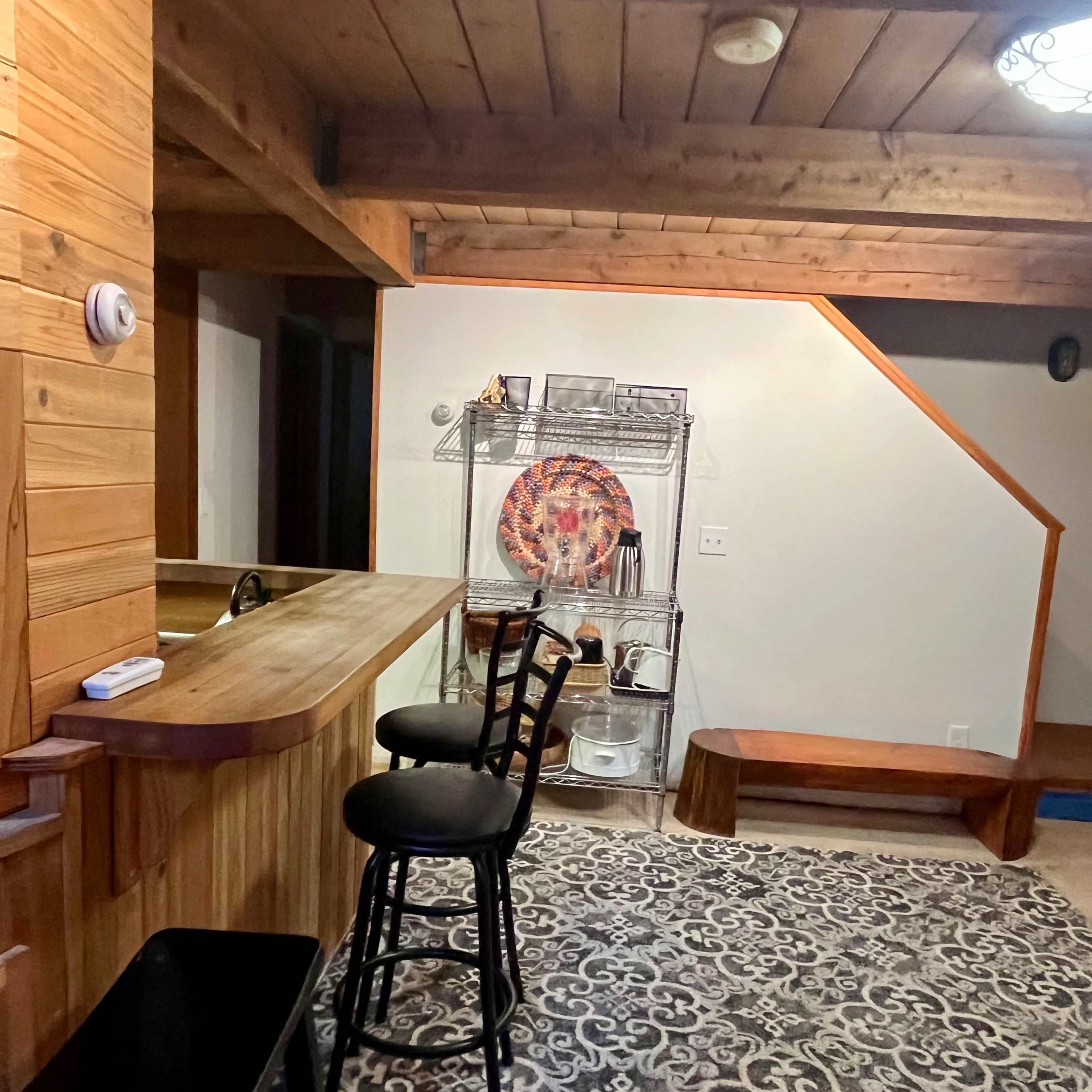 A wooden interior with a bar area, two stools, and a staircase in the background, accompanied by a patterned rug on the floor.