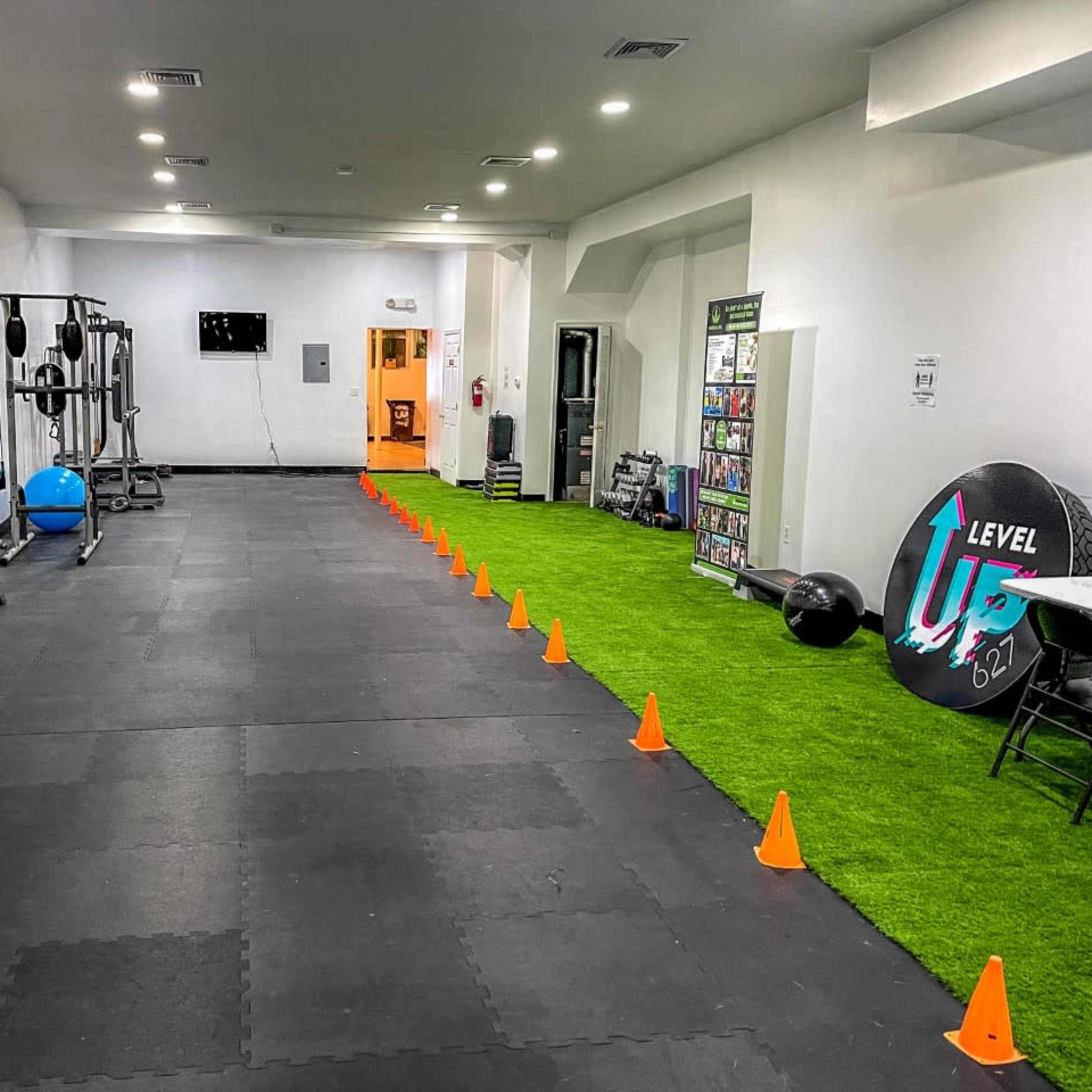 A gym interior with various exercise equipment on one side and a row of orange cones set on artificial grass along the floor.
