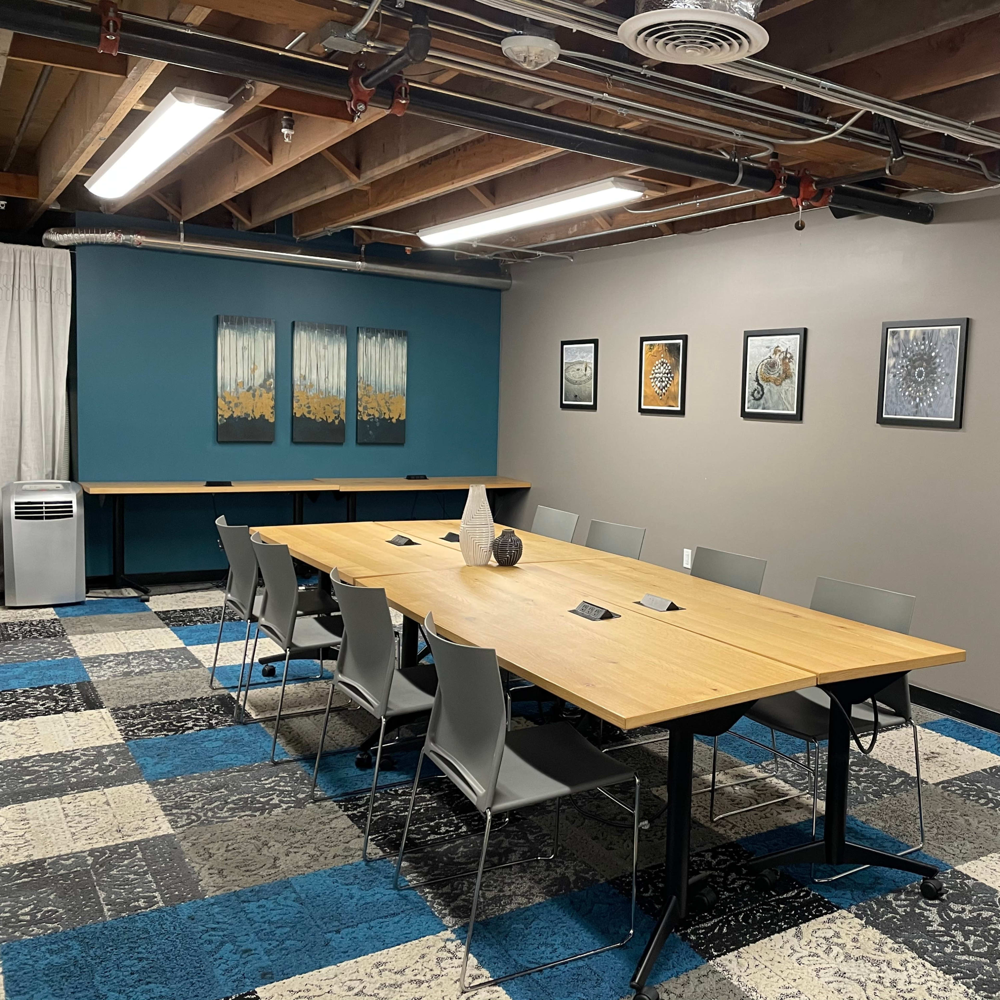 A modern conference room with a long wooden table surrounded by gray chairs, decorated with framed artwork on the walls and featuring a large air conditioning unit in the corner.