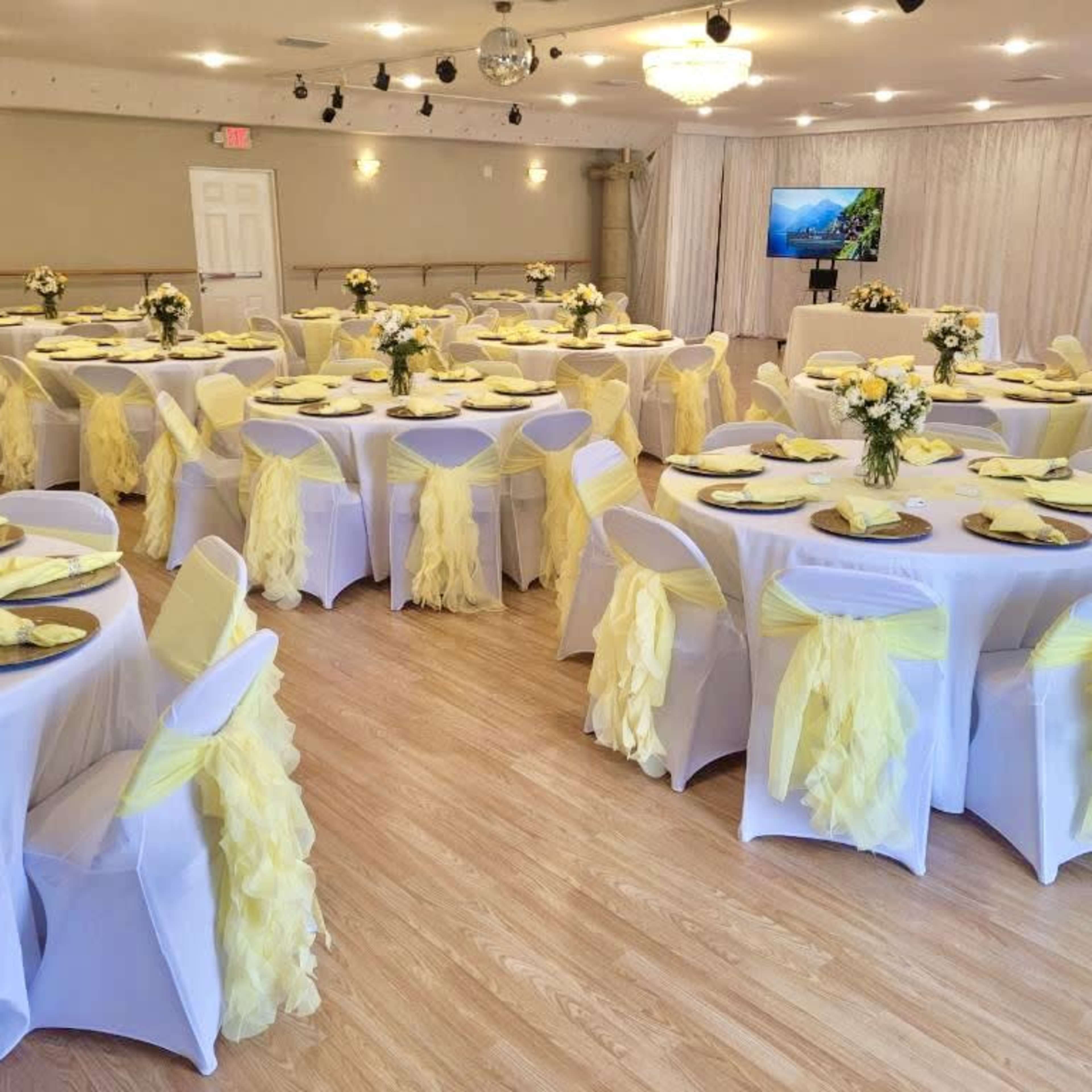 A banquet hall set up for an event with round tables covered in white tablecloths and decorated with yellow-tinted centerpieces, surrounded by chairs draped in yellow fabric.