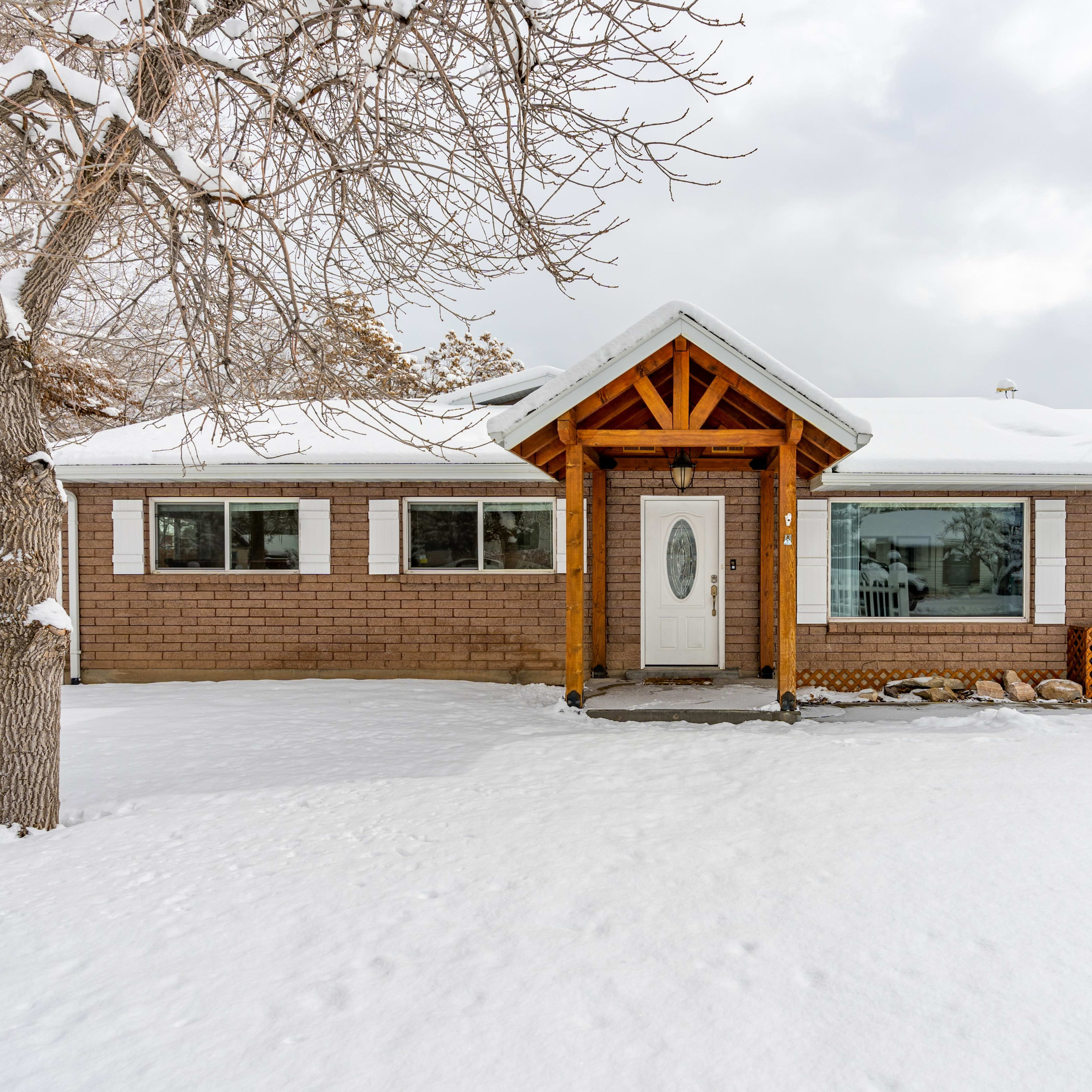 A single-story brick house with a wooden porch structure is surrounded by snow, featuring large front windows and an attached garage.
