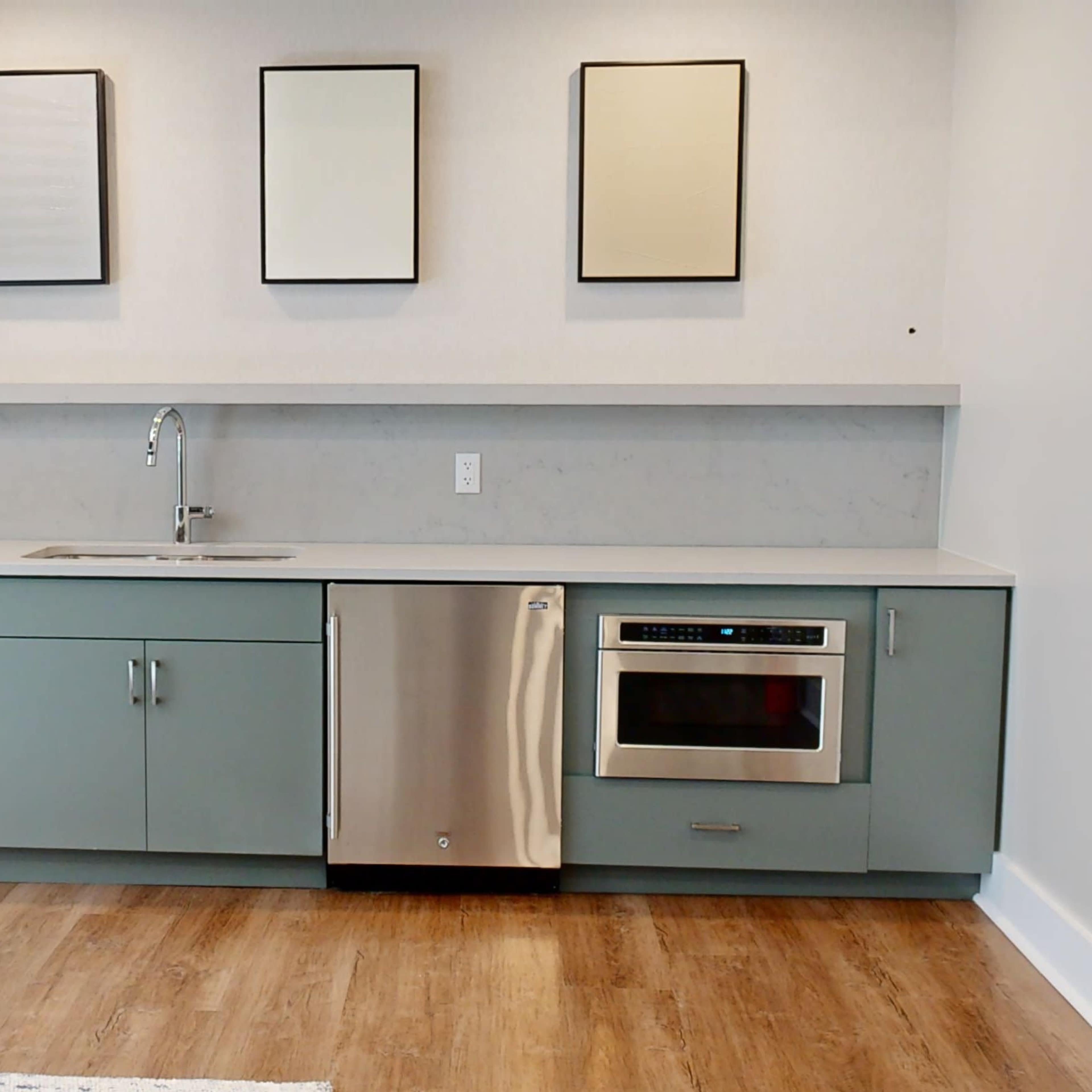 A modern kitchen with light green cabinets, a stainless steel dishwasher and microwave, and three empty framed pictures on the wall above the countertop.