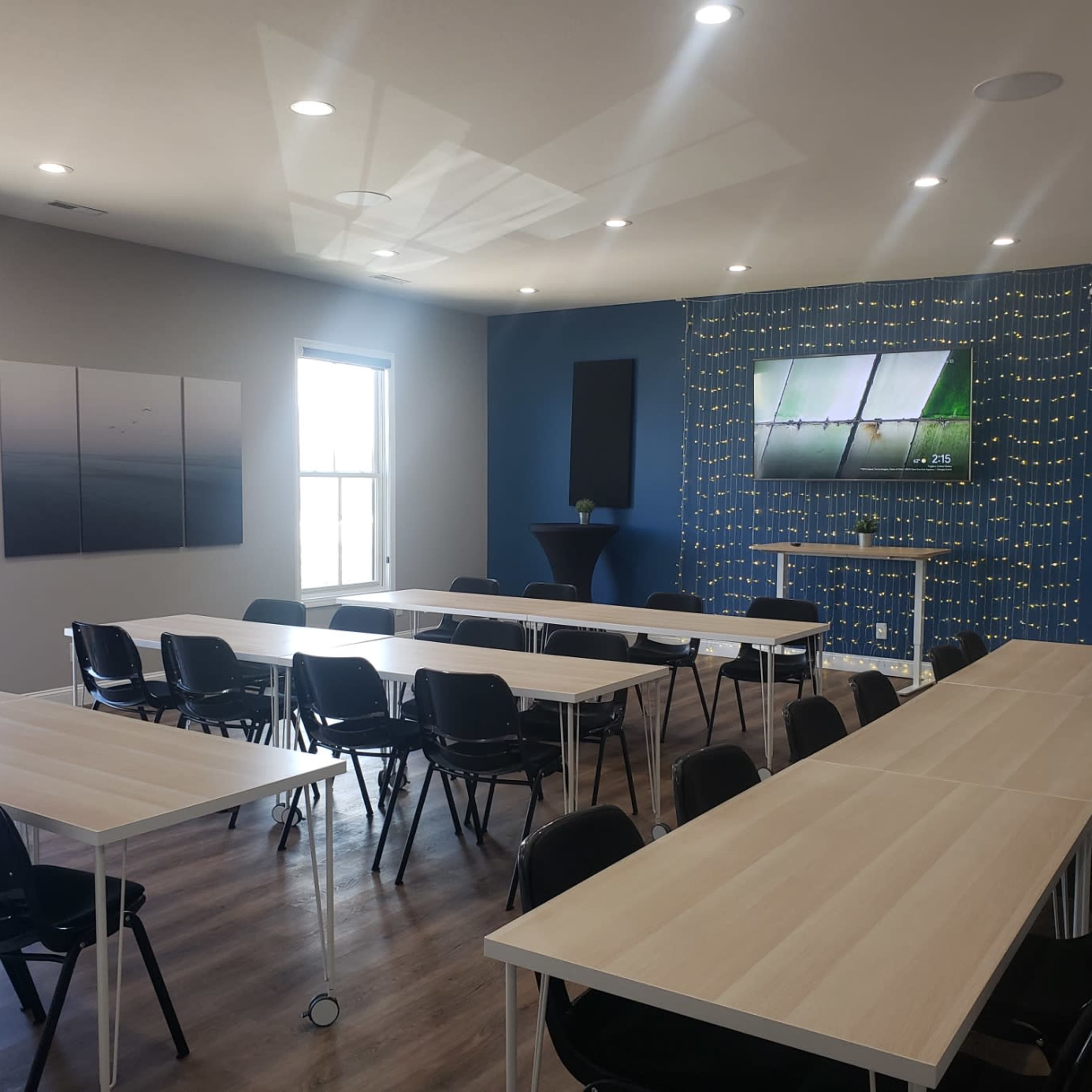 A modern meeting room with multiple tables arranged in a U-shape, black chairs, and a large screen mounted on a blue accent wall adorned with decorative lighting.