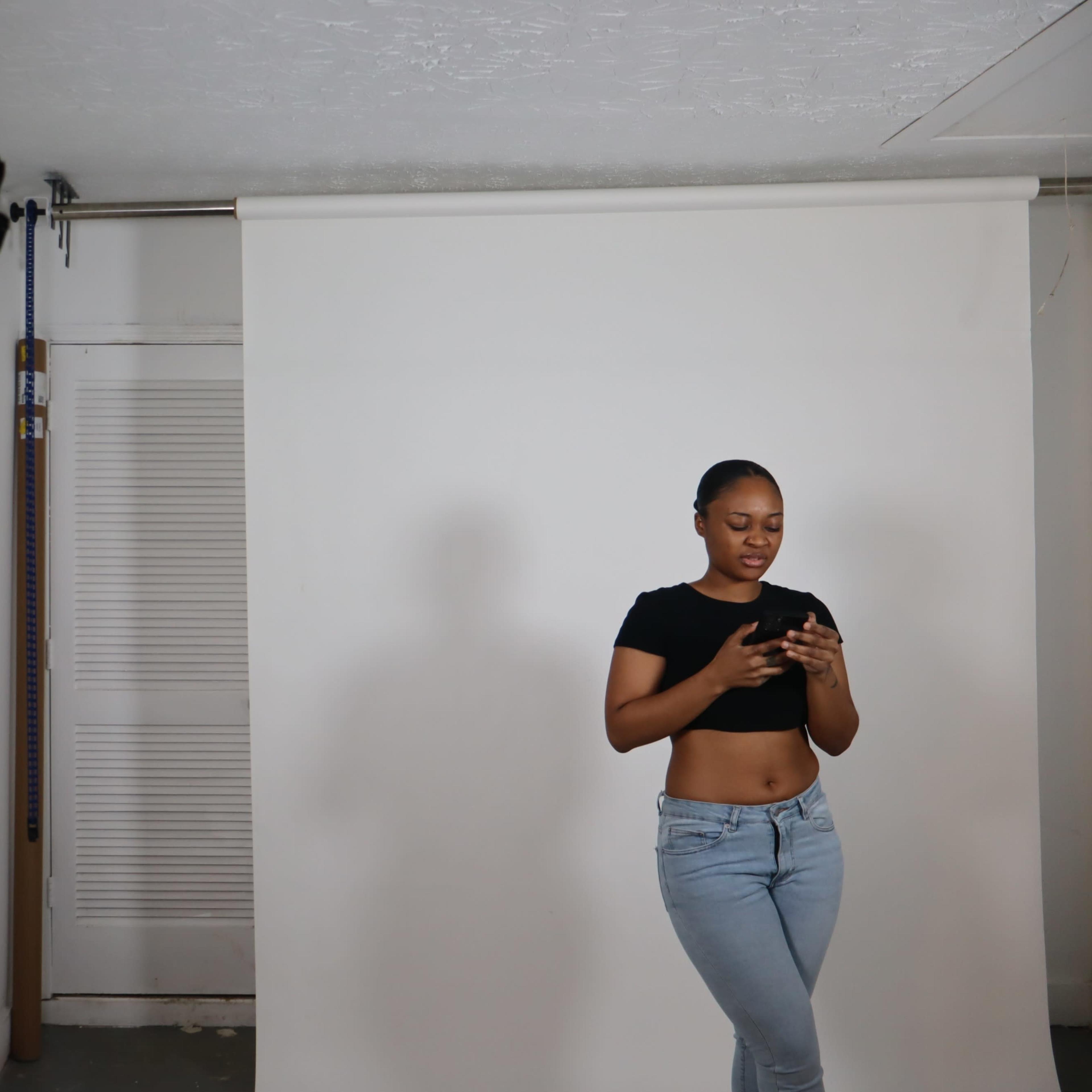A woman in a black crop top and blue jeans stands in a photography studio, looking at her phone against a plain white backdrop.