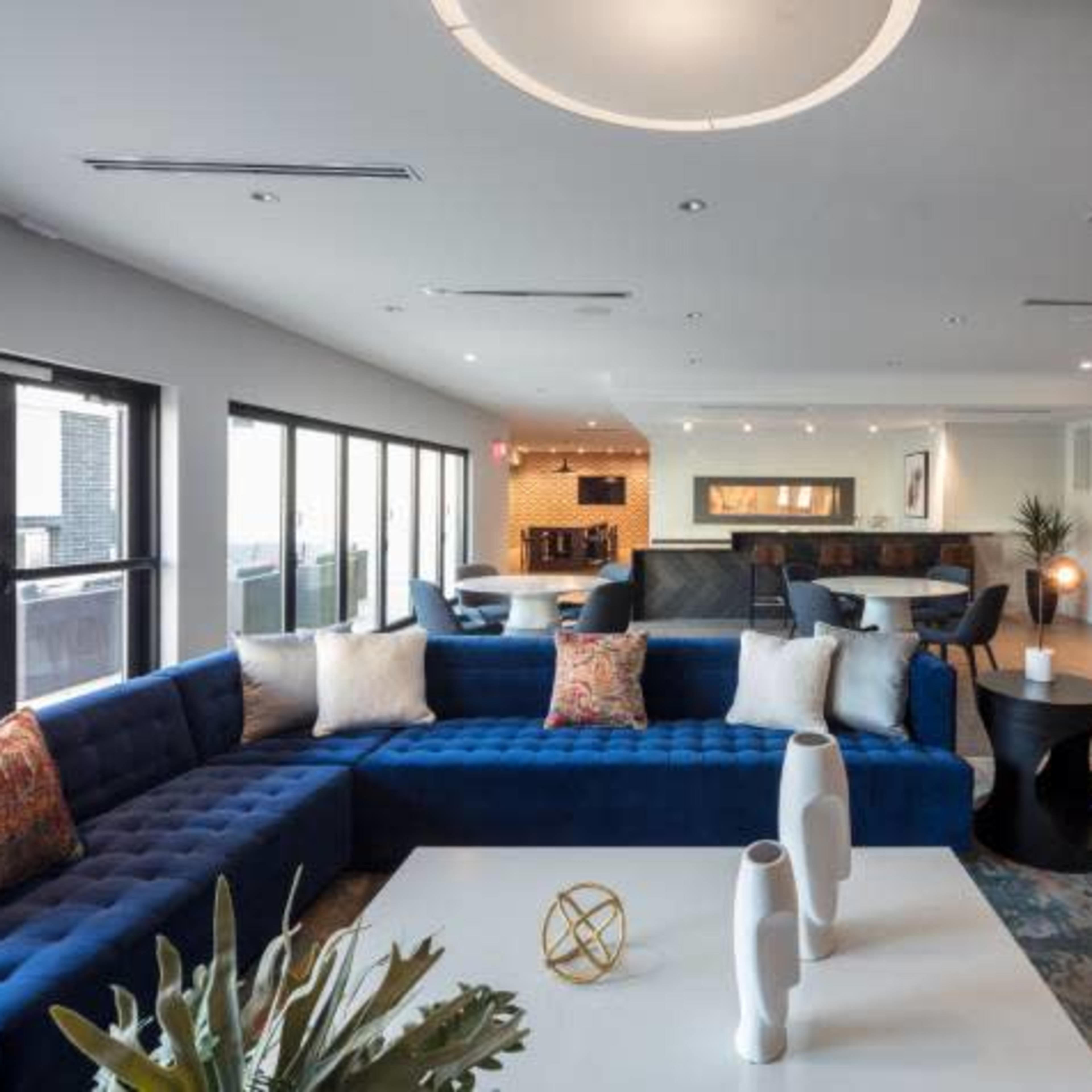 A modern lounge area featuring a blue sectional sofa, white coffee table, and various seating arrangements with large windows providing natural light.