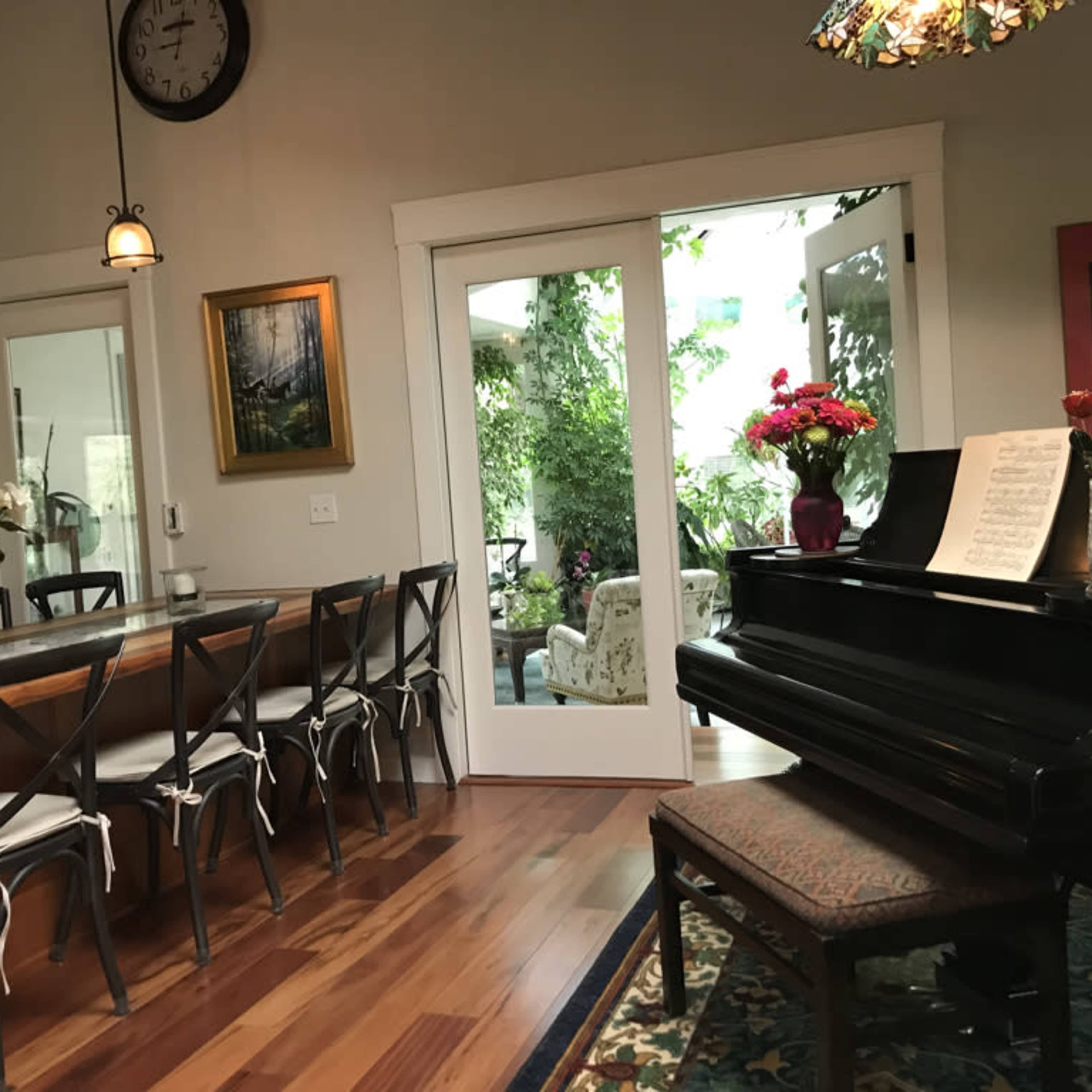 A dining area with a large wooden table and chairs adjacent to a grand piano, with glass doors that open to a garden space.