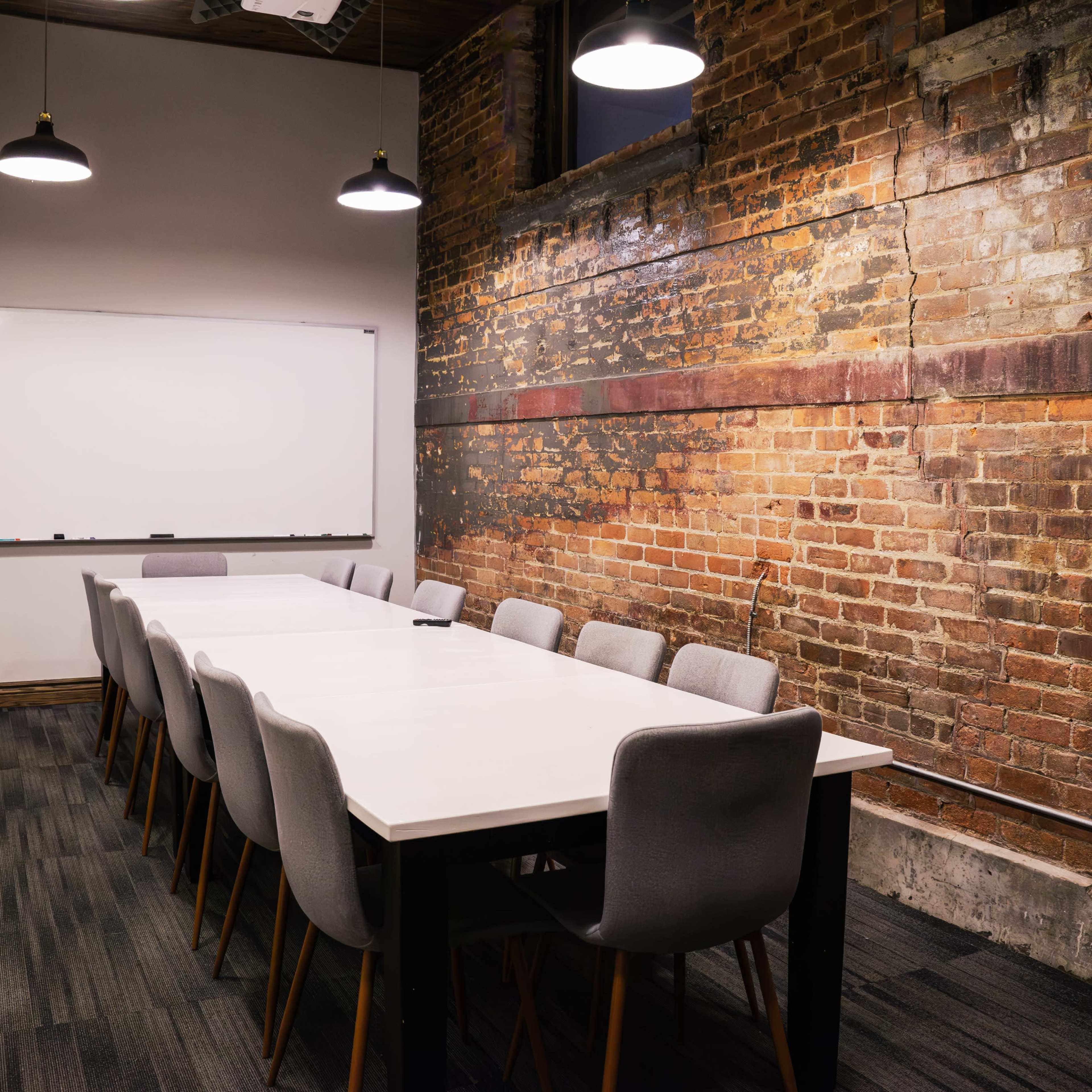 A conference room features a long white table surrounded by gray chairs, against a backdrop of exposed brick walls.