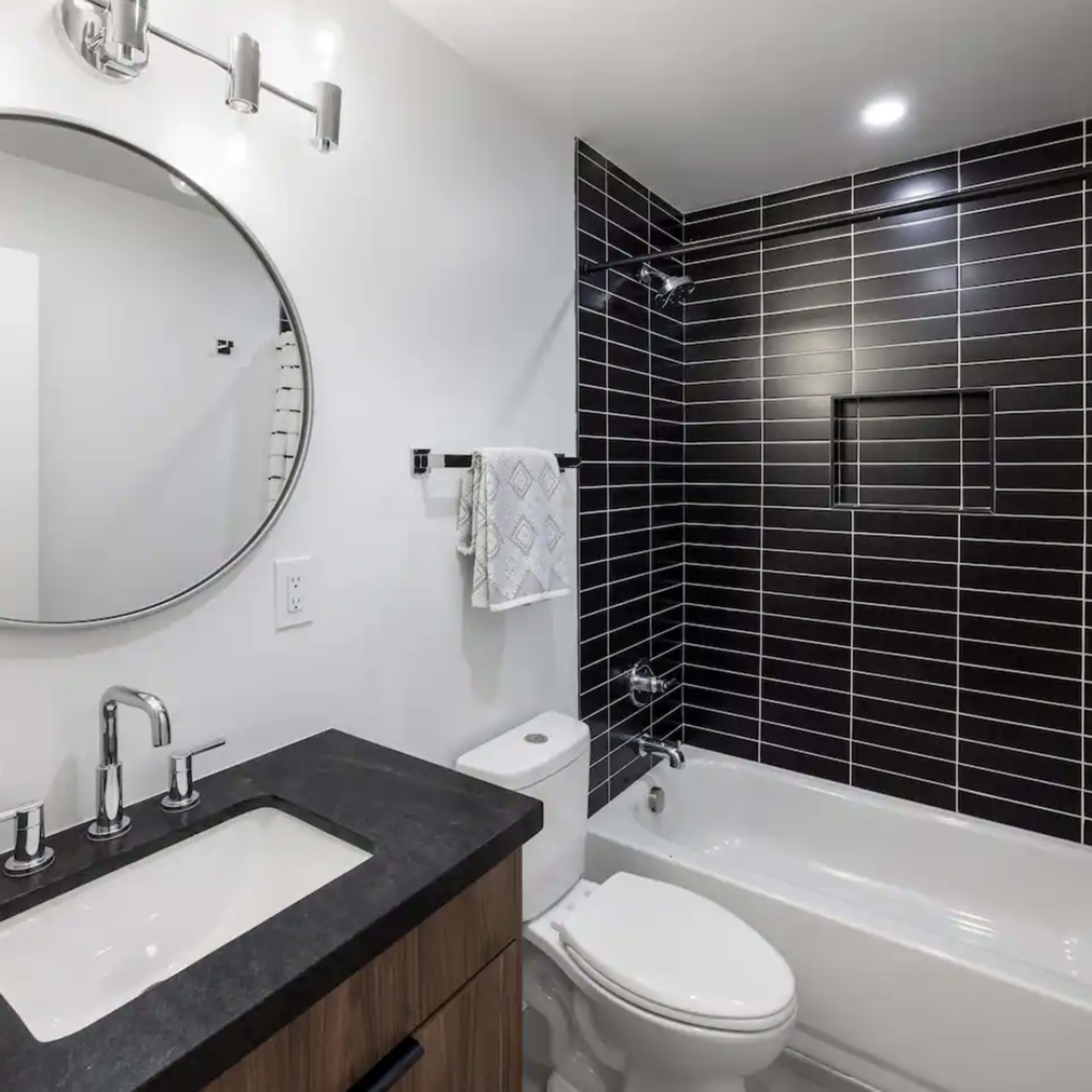 A modern bathroom featuring a round mirror, a dark tiled wall, a white bathtub, and a sink with a dark countertop.