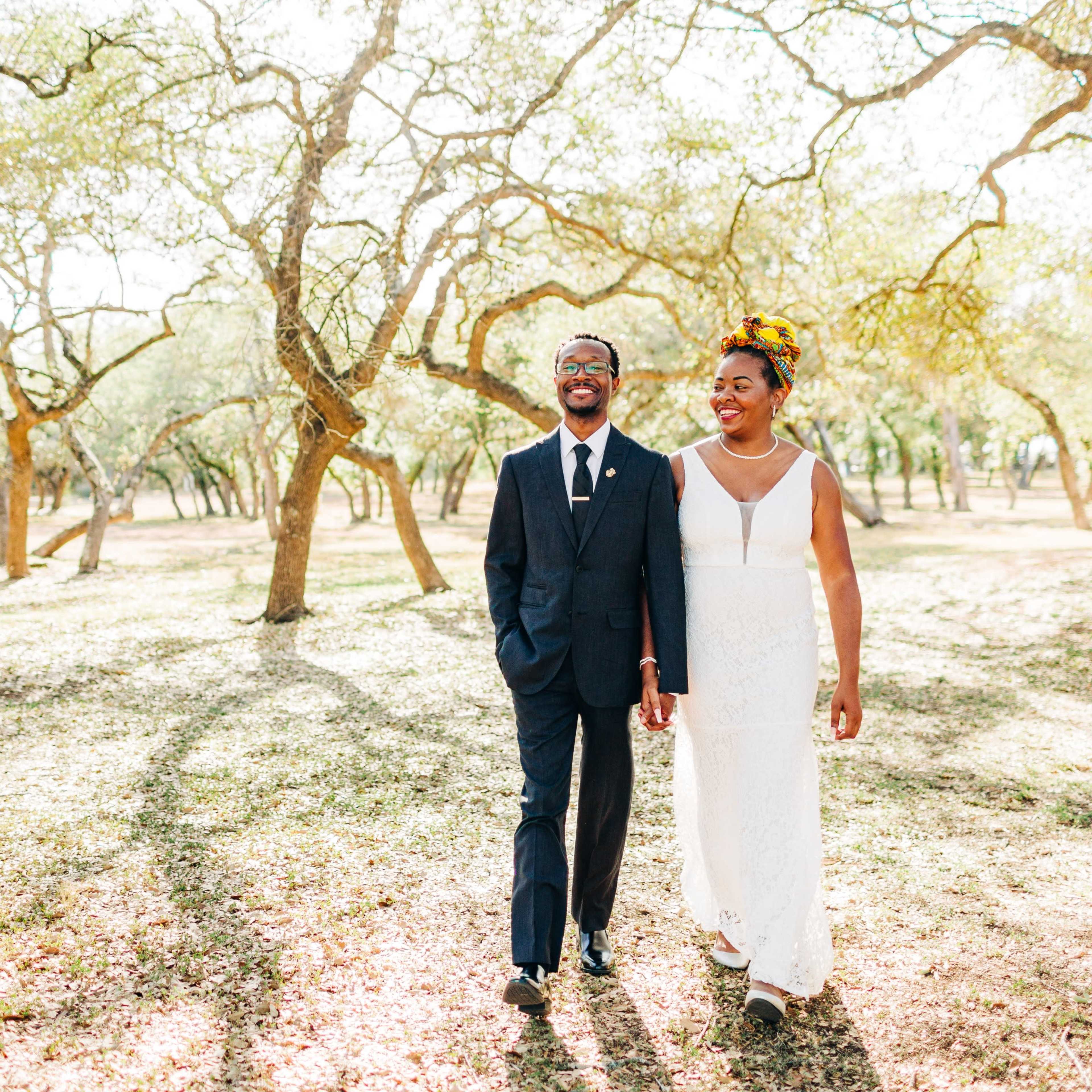 A couple walks hand in hand along a sunlit path surrounded by trees in a park setting.