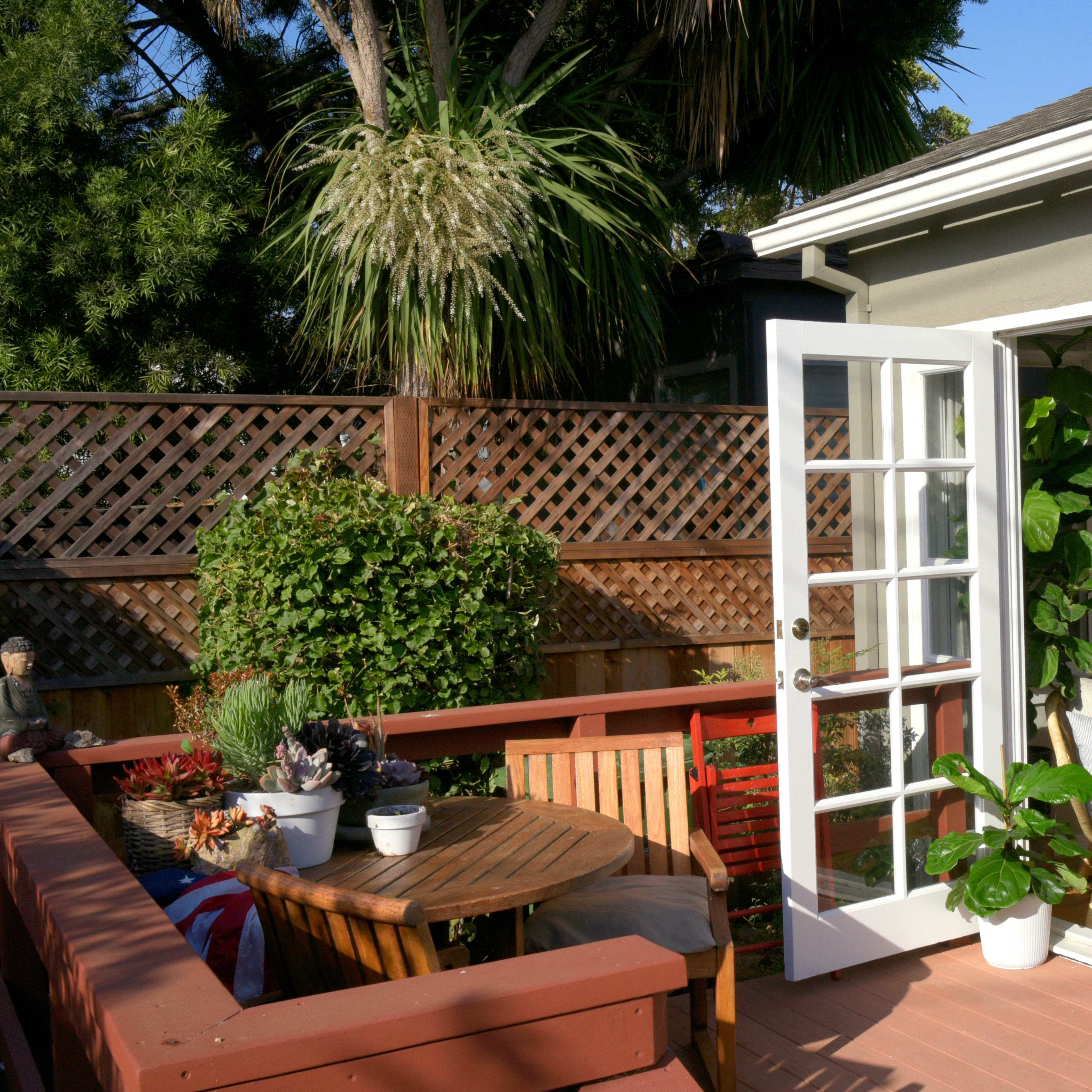 A deck with a round table and chairs is adjacent to a house with open French doors, leading to a cozy indoor space filled with plants.