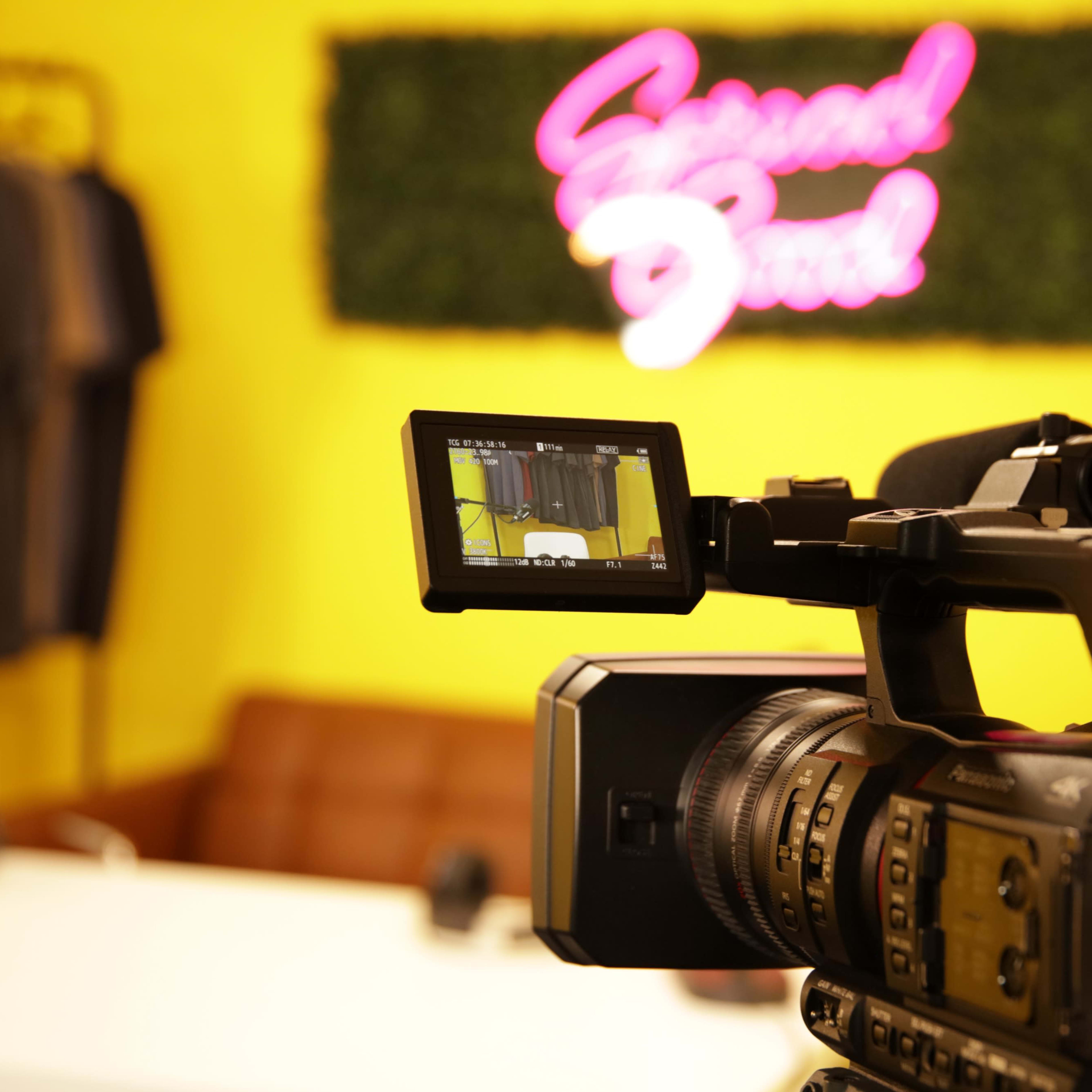 A video camera is positioned in front of a brightly colored wall featuring a neon sign that reads "Second Soul," with clothing arranged in the background.