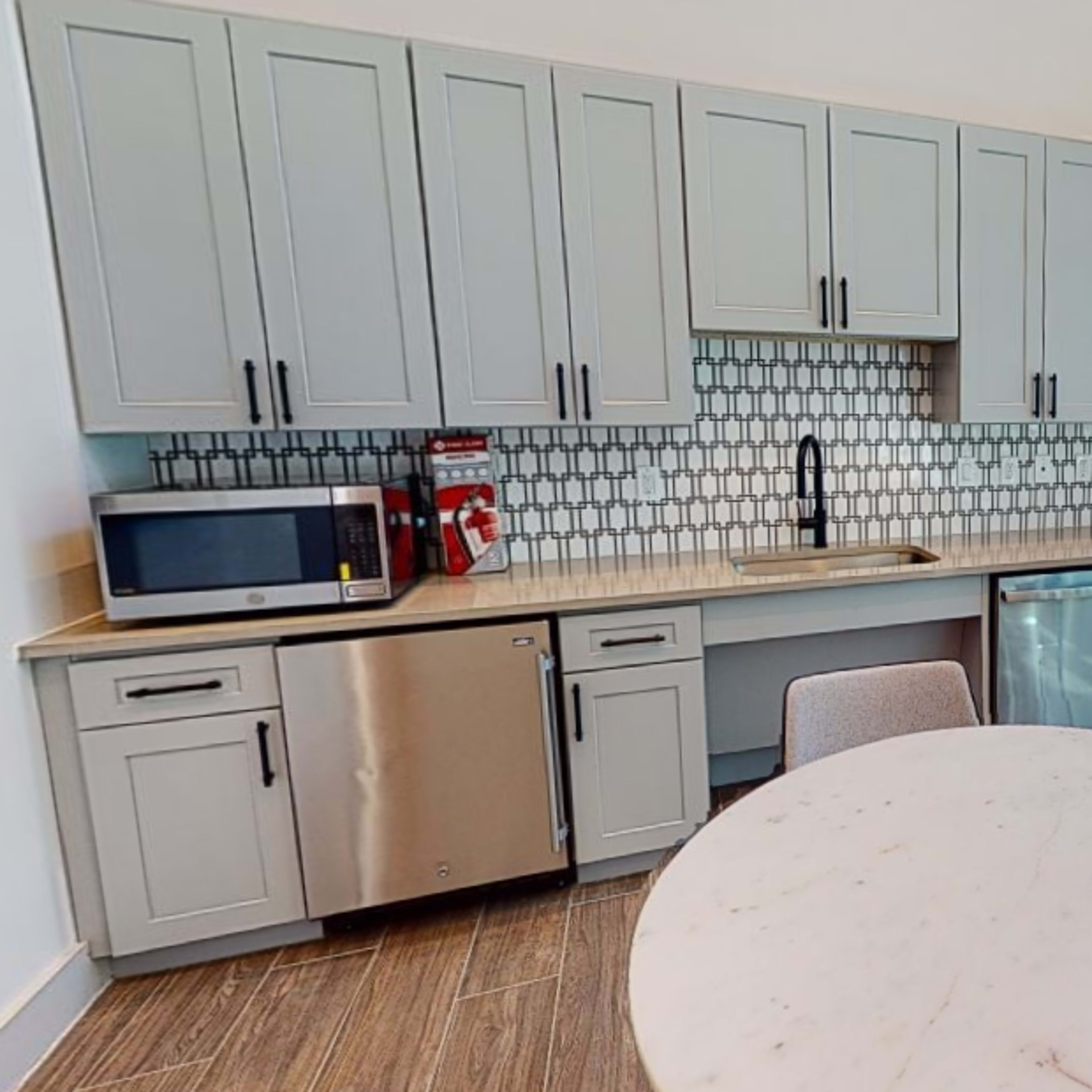 A modern kitchen space featuring gray cabinets, a microwave, a dishwasher, a marble-topped table, and large windows providing natural light.
