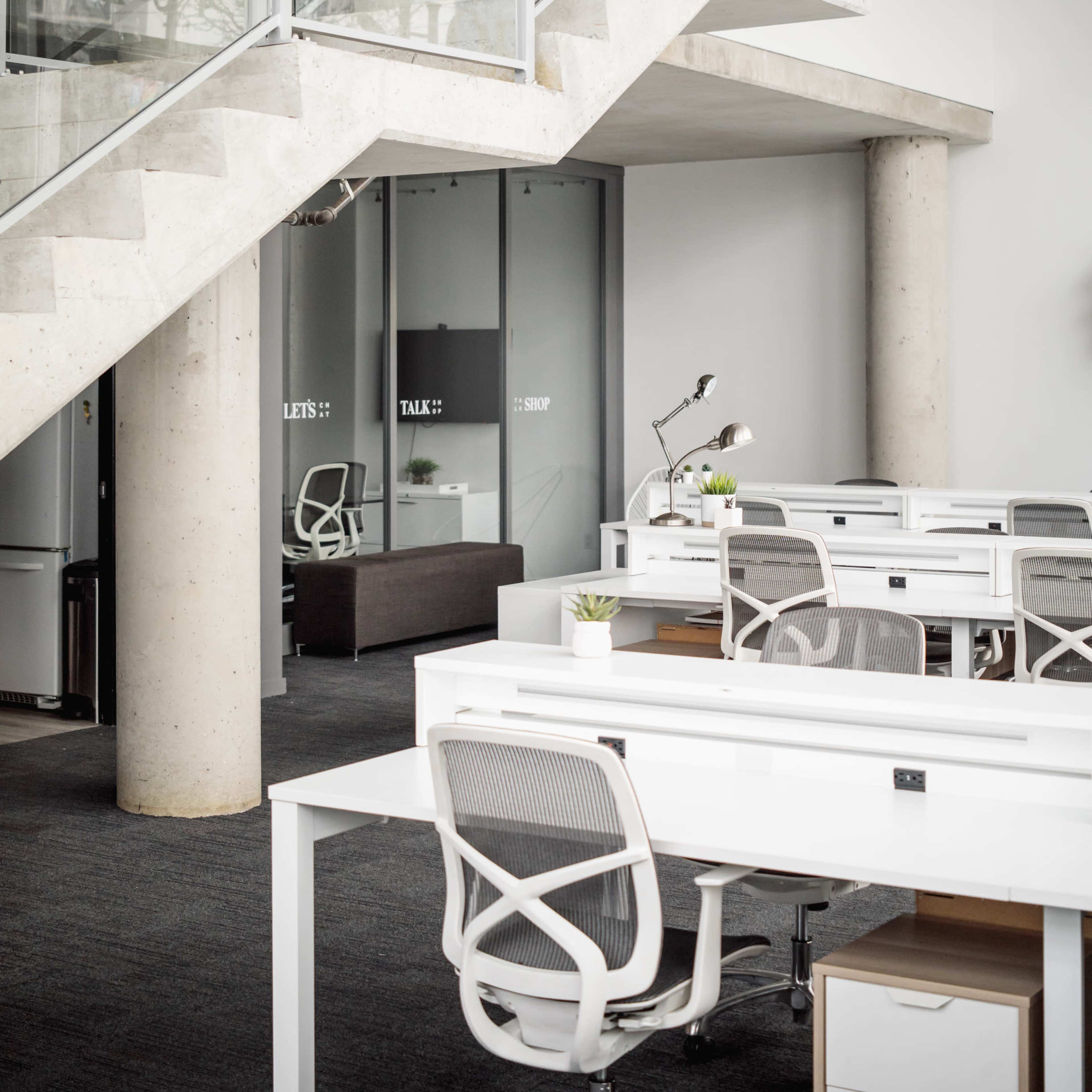 A modern office space featuring a staircase, glass partitions, and several white desks arranged throughout the area.