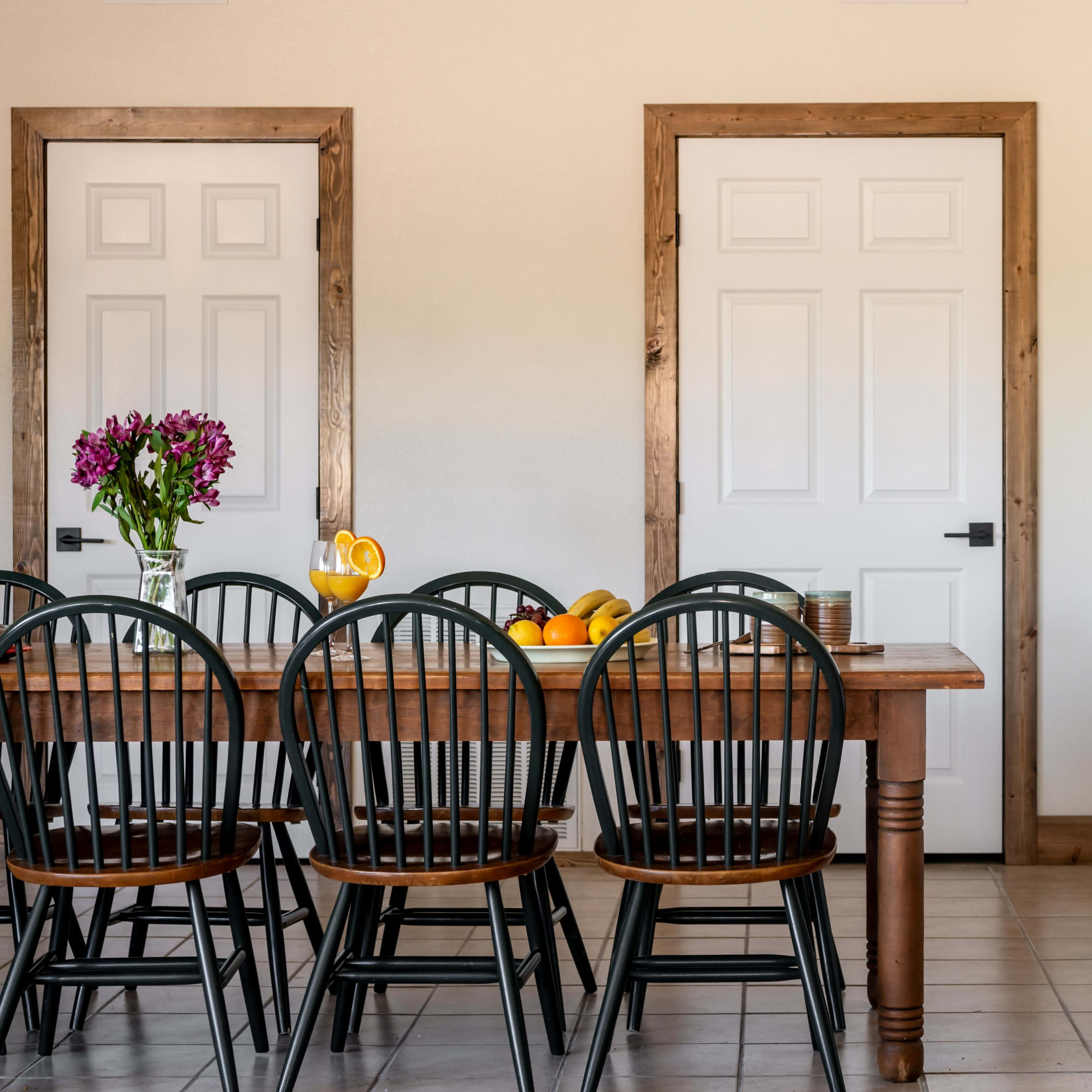 A wooden table with black chairs is set against a wall with two mirrors and a closed white door, alongside a wooden cabinet with decorative items.