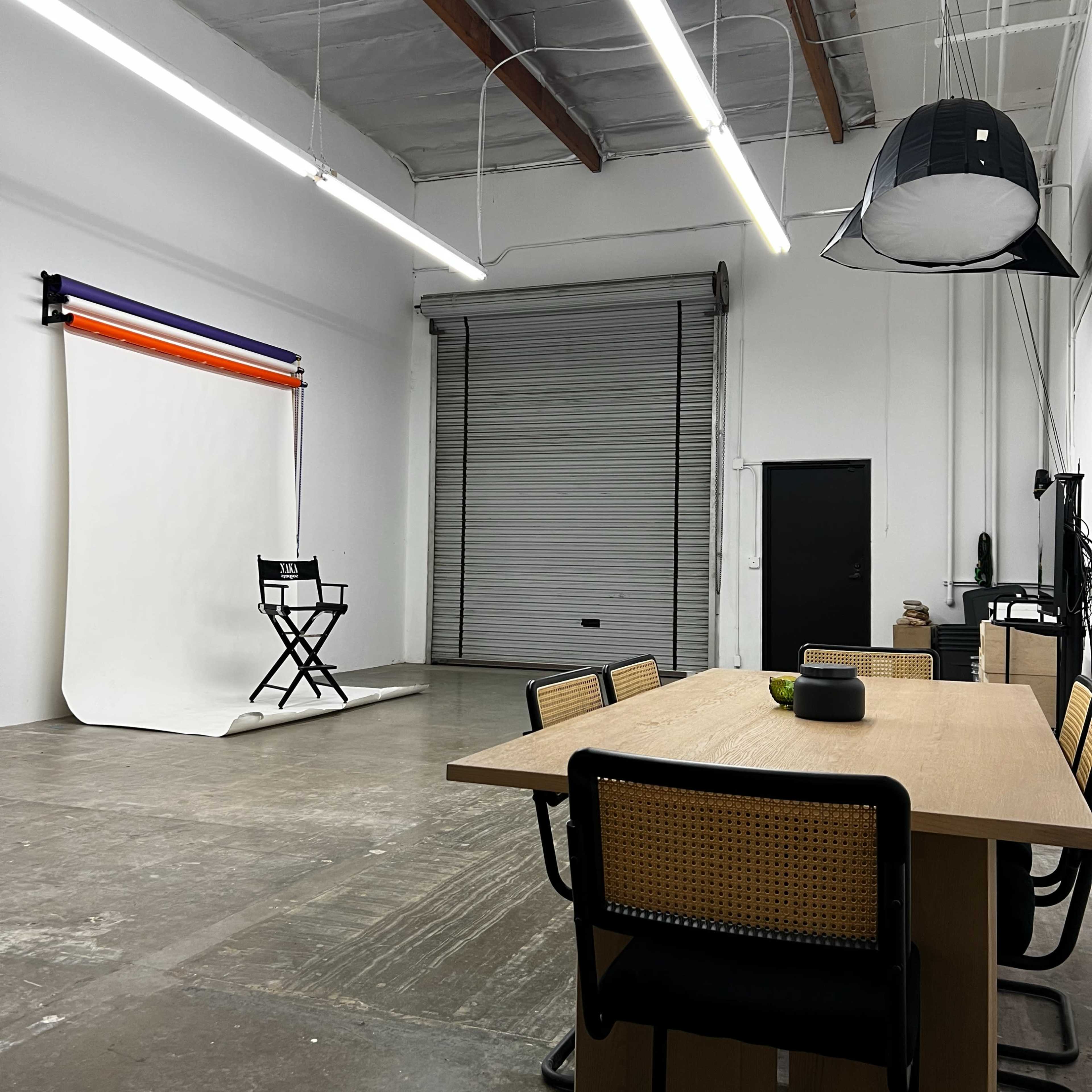 A modern studio space features a large wooden table with black chairs, a roll-up garage door, and a backdrop setup for photography.