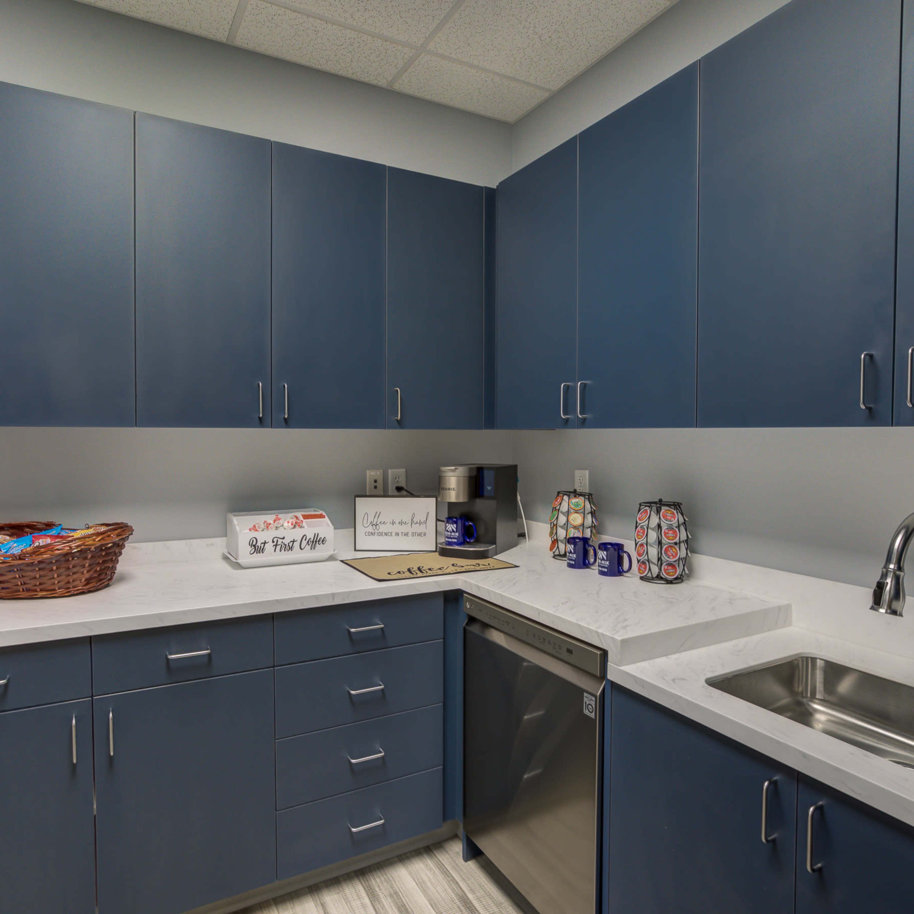 A modern kitchen with blue cabinets, a stainless steel dishwasher, a sink, and a countertop displaying a basket and decorative mugs.