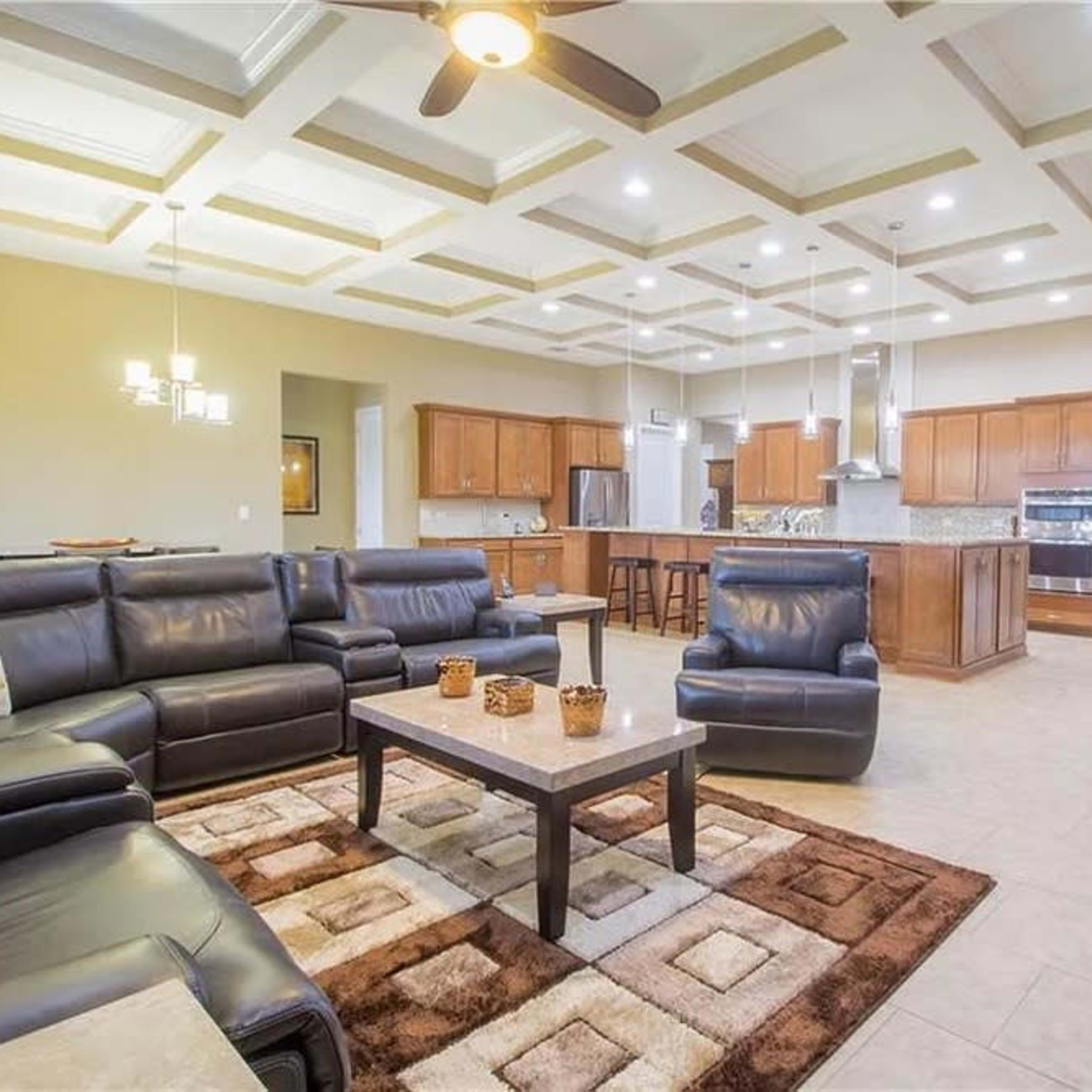 A spacious, modern living room with a coffered ceiling, dark leather furniture, and an open layout connecting to a kitchen area.