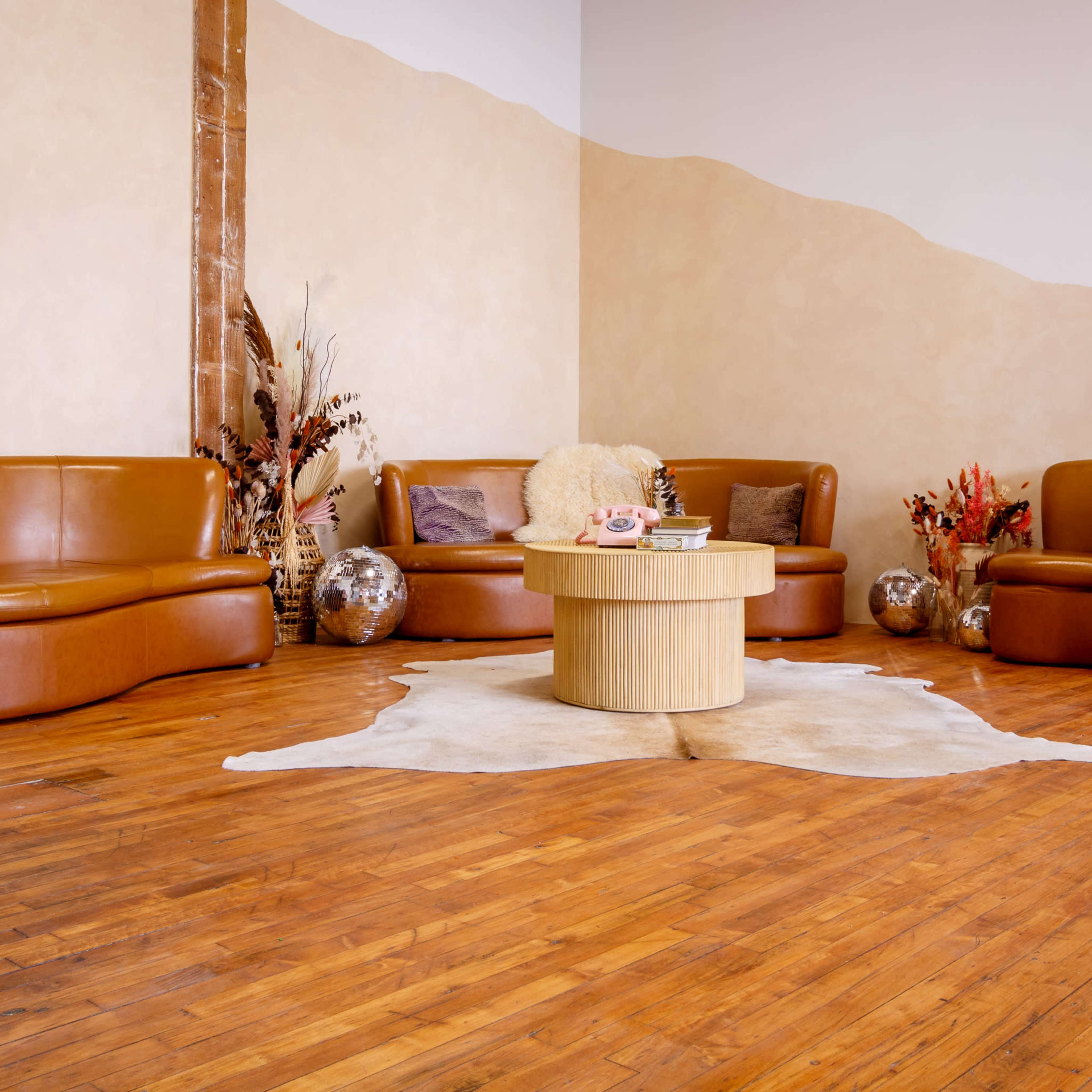 A circular wooden coffee table surrounded by curved brown leather couches on a wooden floor, with decorative elements like vases and plants in the background.