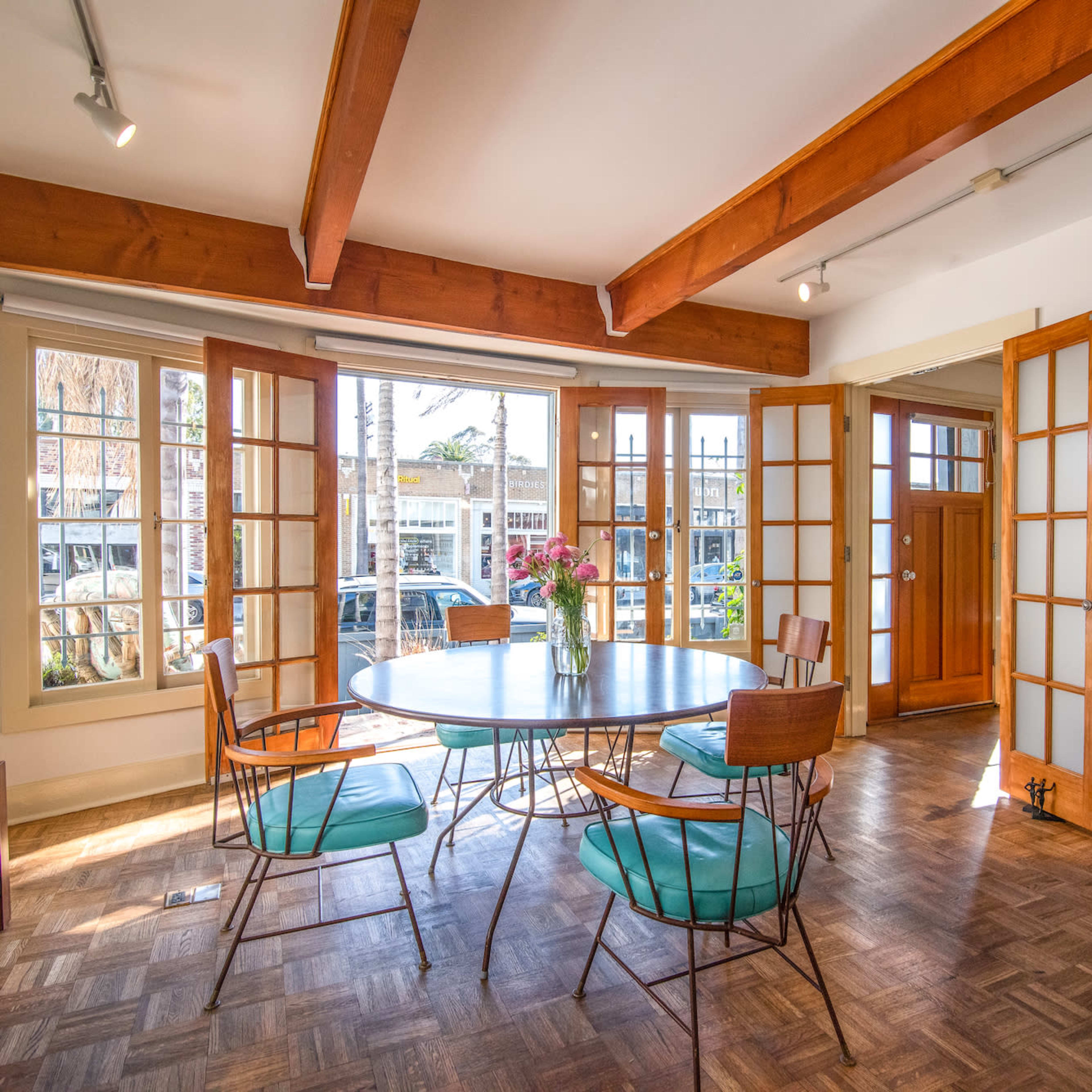 A dining area features a round glass table surrounded by wooden chairs, with multiple large windows letting in natural light.