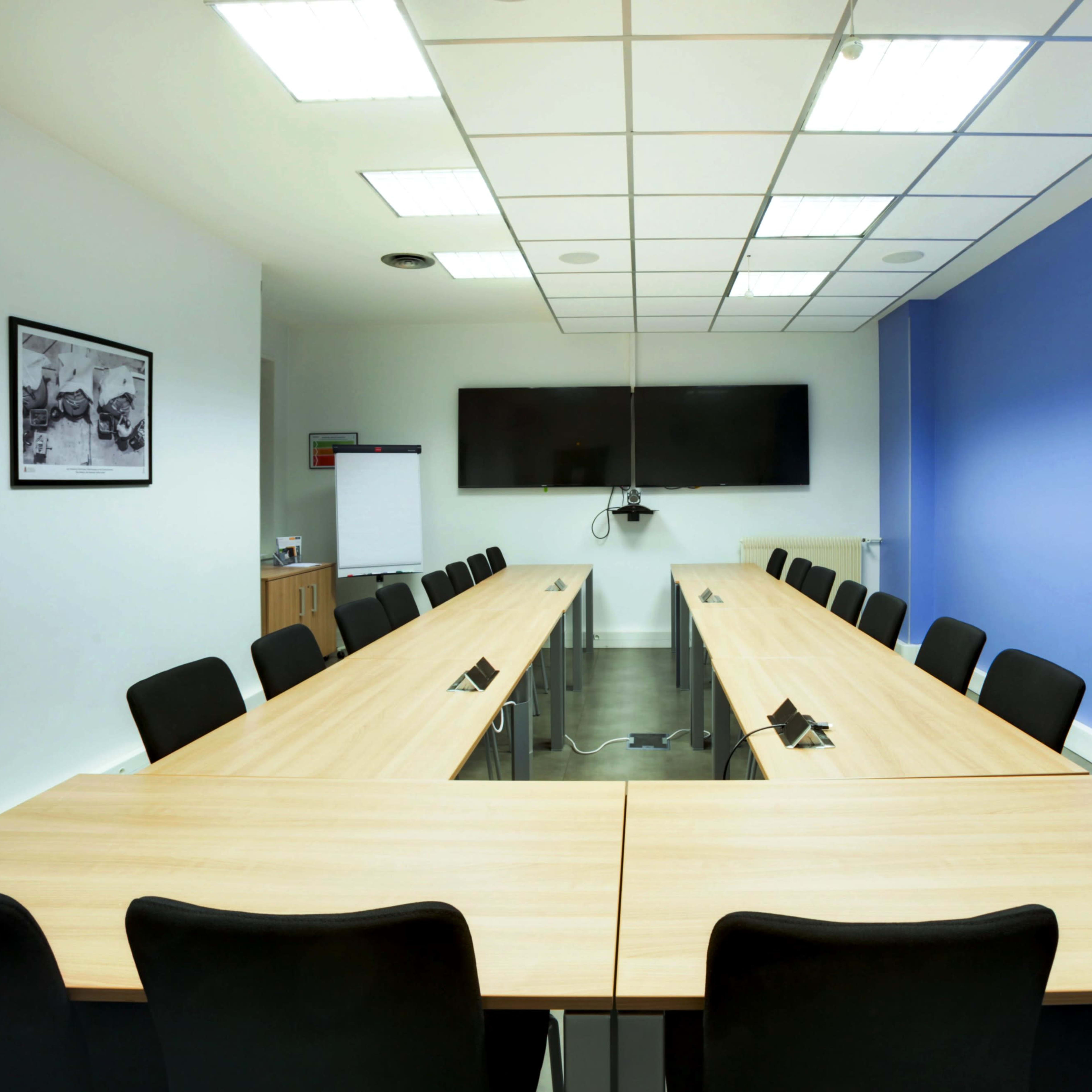A conference room features two long wooden tables arranged in a U-shape, surrounded by black chairs, with a large screen mounted on the wall and a blue accent wall.