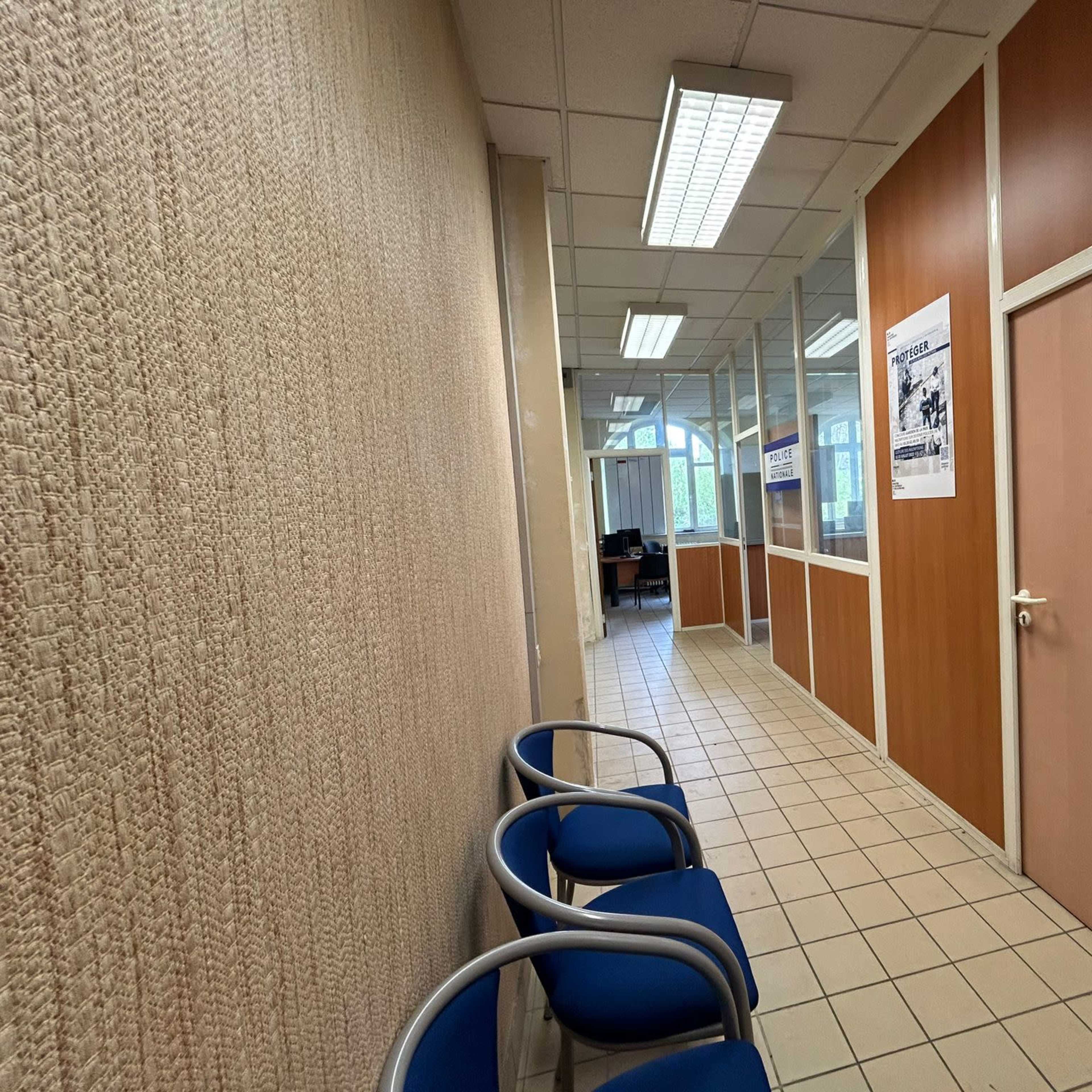 A corridor with beige walls, tiled flooring, and several blue chairs lined up against one side, leading to a door at the end.