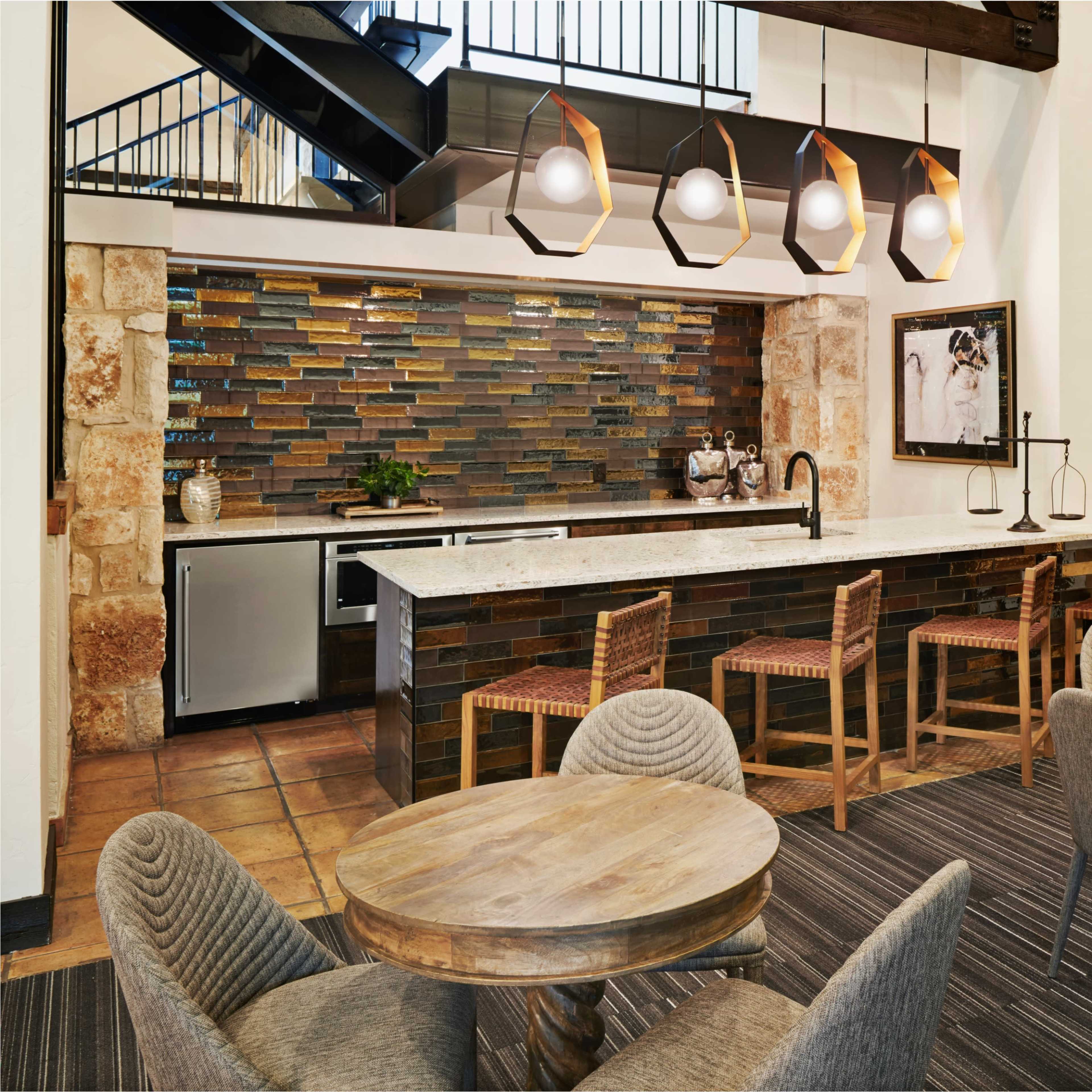 A modern kitchen area with a granite countertop, bar stools, and a colorful tile backsplash, surrounded by dining tables and decorative lighting fixtures.