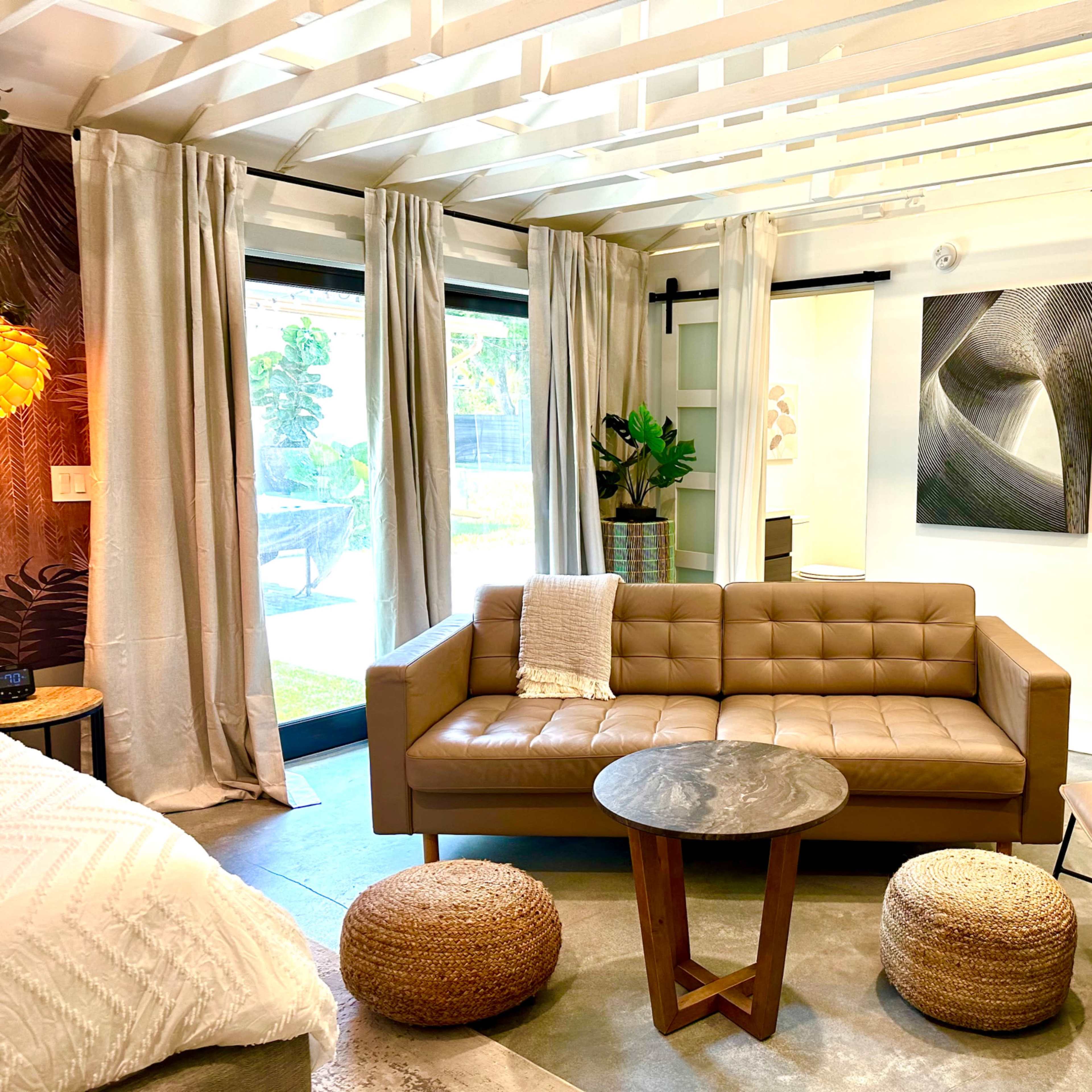 A modern interior living space featuring a brown leather sofa, a round coffee table, woven poufs, and large windows with sheer curtains.