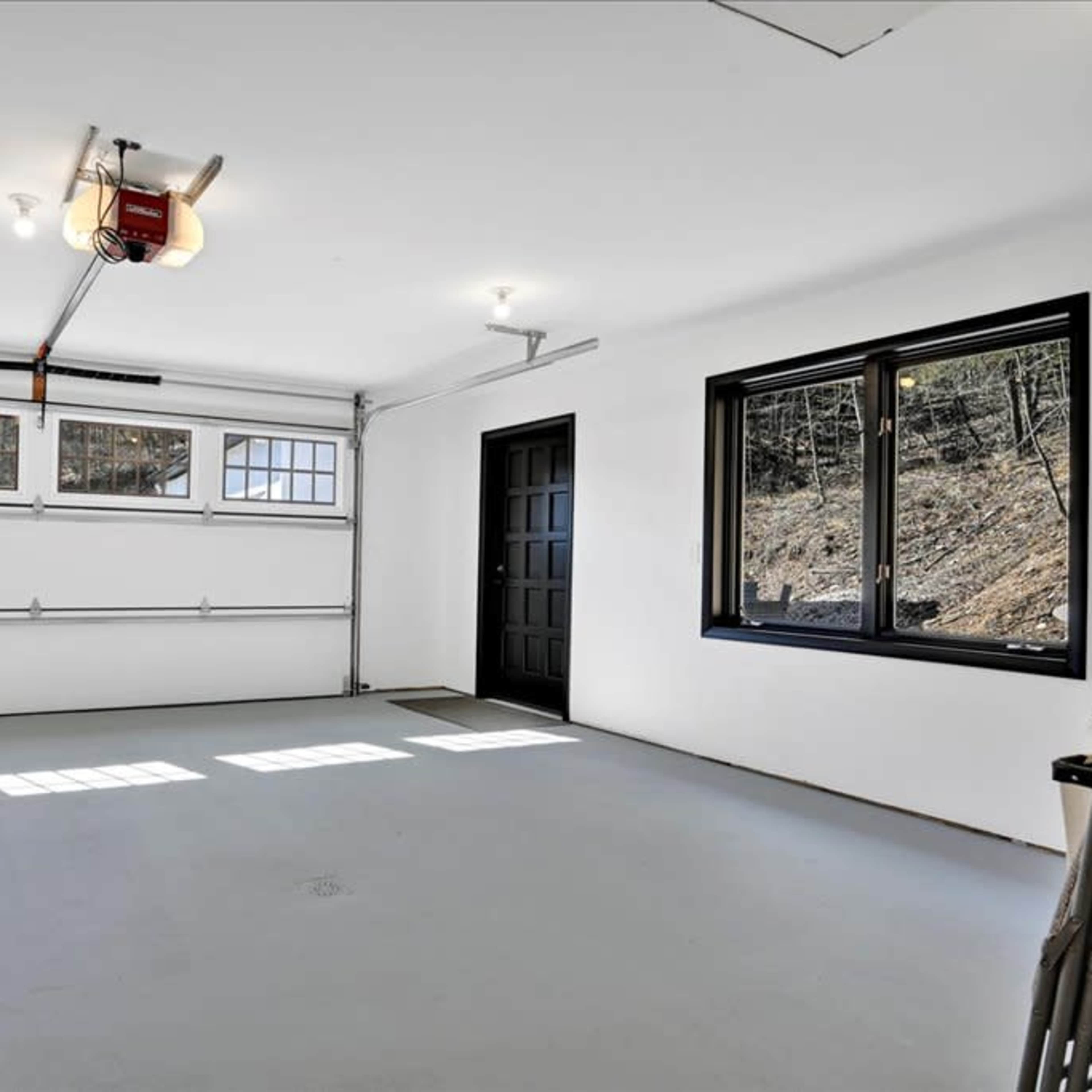 A spacious, empty garage with a large overhead door, a side entrance door, a window, and a trash can.