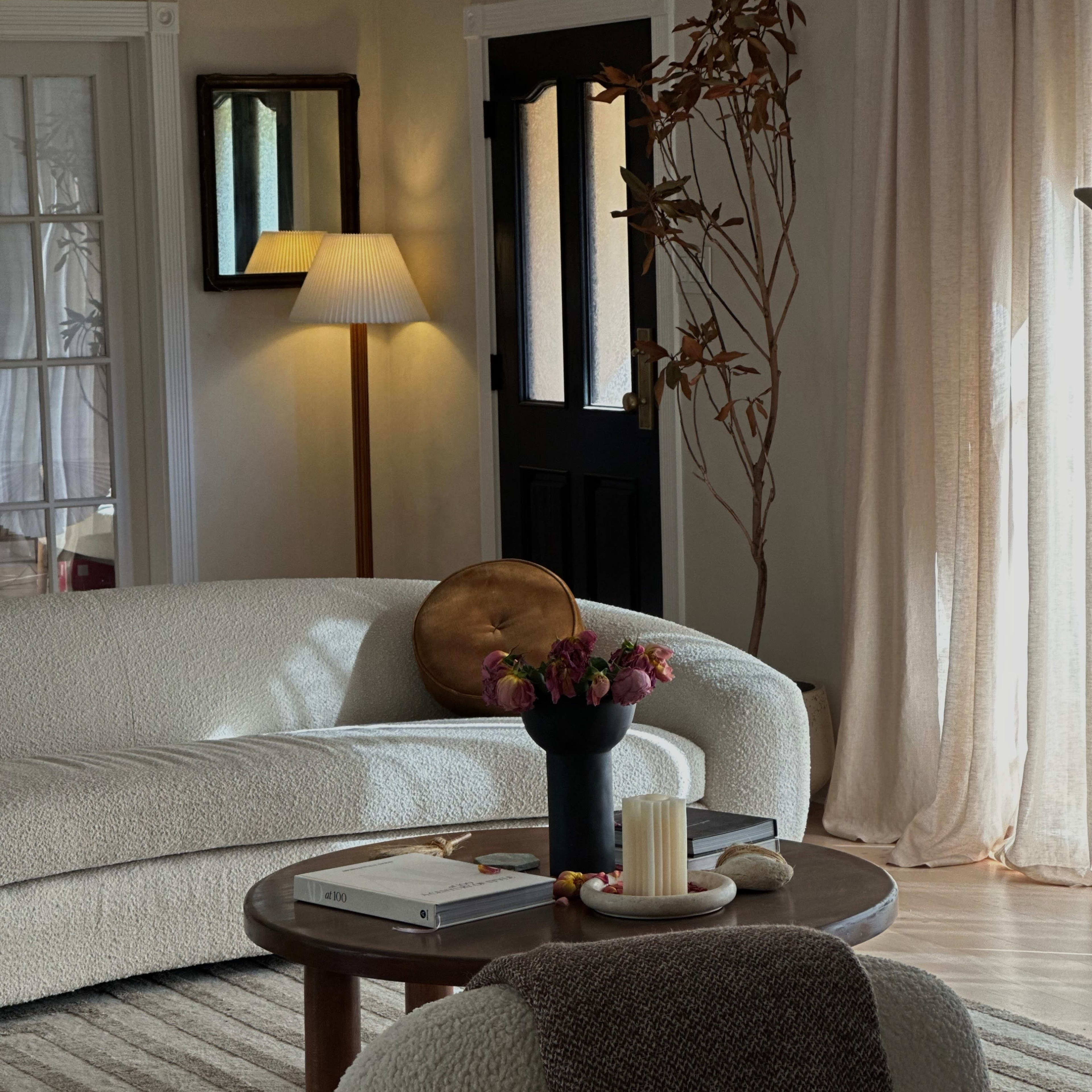 A cozy living room featuring a white, textured sofa, a round wooden table with a vase of flowers, and a tall lamp beside a dark door.