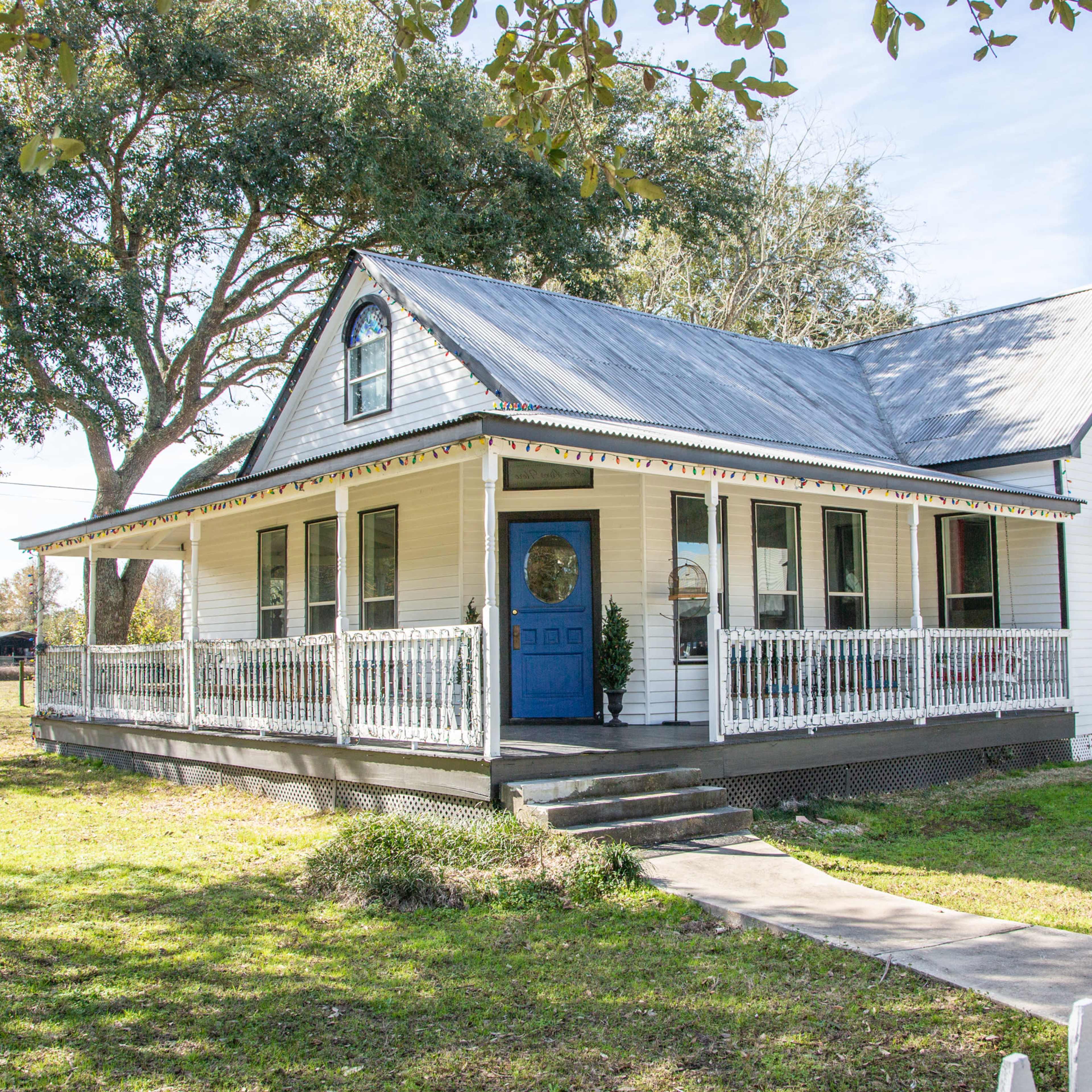 A white, two-story house with a blue door, surrounded by a green lawn and trees.