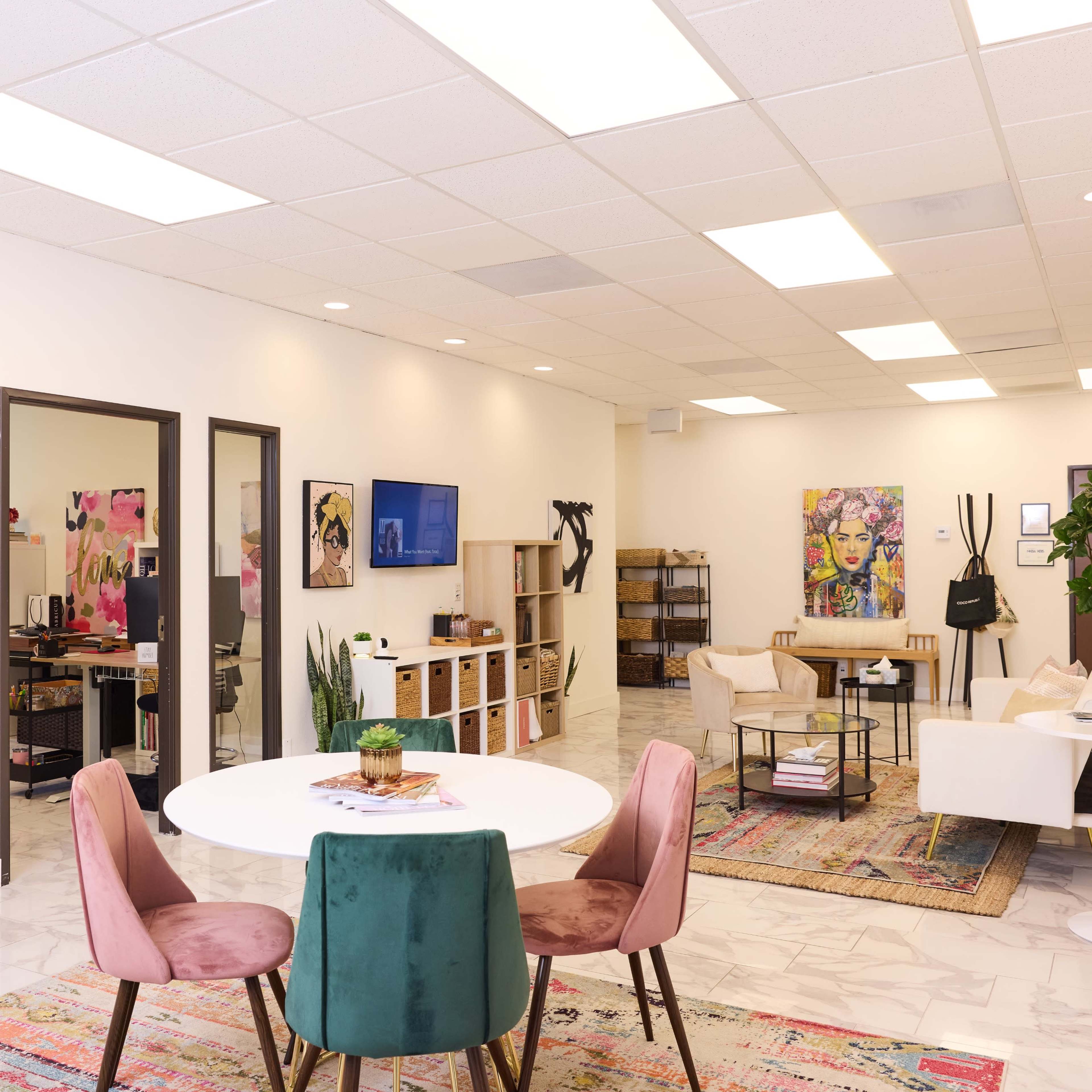 A modern office space features a circular table with pink chairs, large artworks on the walls, and a mix of workstations and seating areas.