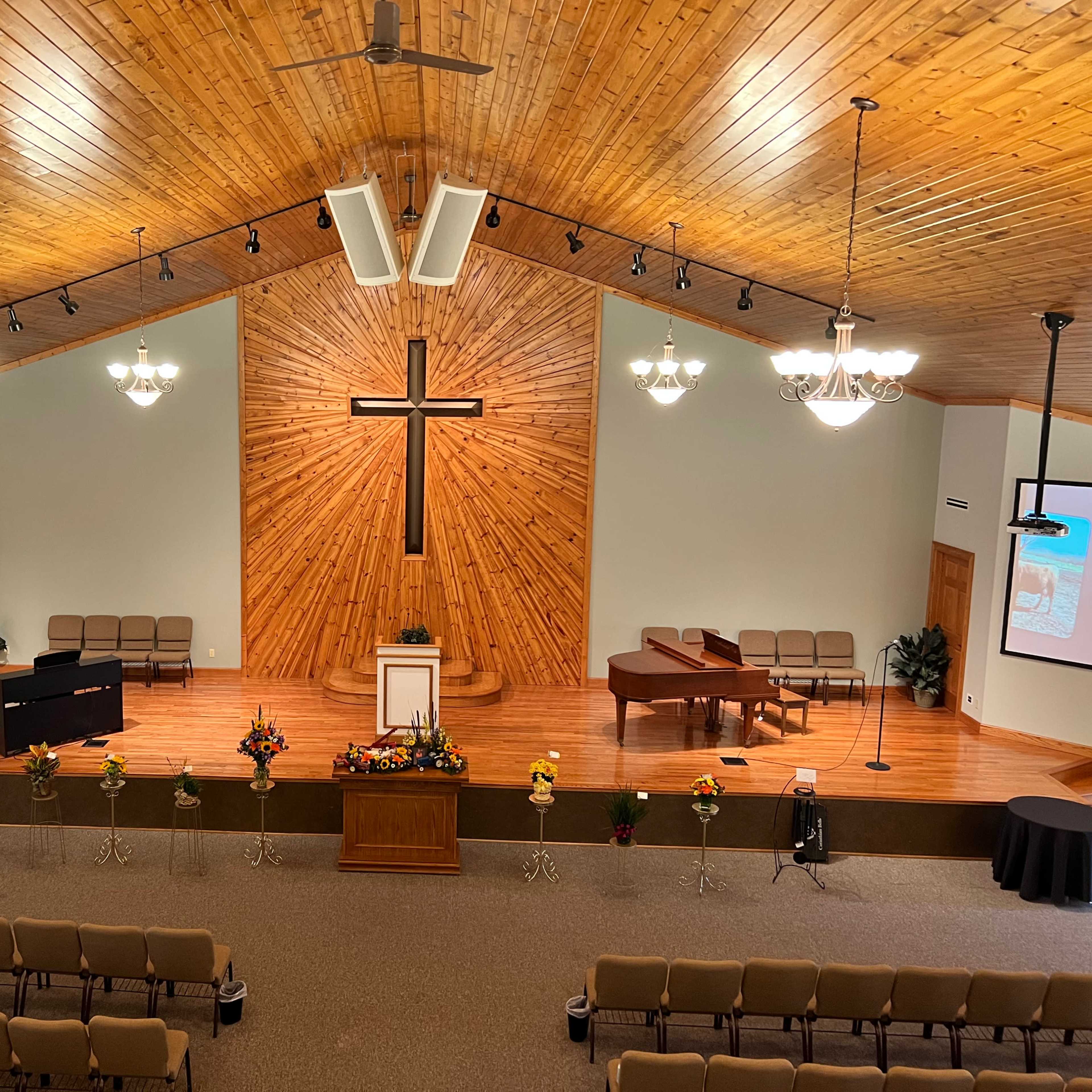 The interior of a church features wooden paneling, a large cross on the wall, a stage with a piano, and rows of chairs arranged for a congregation.